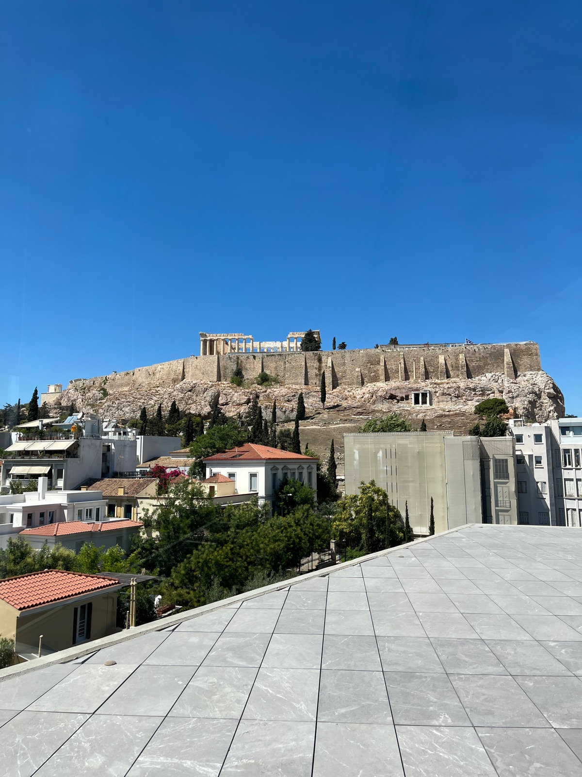 Ο Παρθενώνας και τα τείχη της Ακρόπολης ιδωμένα από τη βεράντα του μουσείου κάτω από λαμπερό γαλάζιο ουρανό, στέγες και δέντρα ανάμεσα