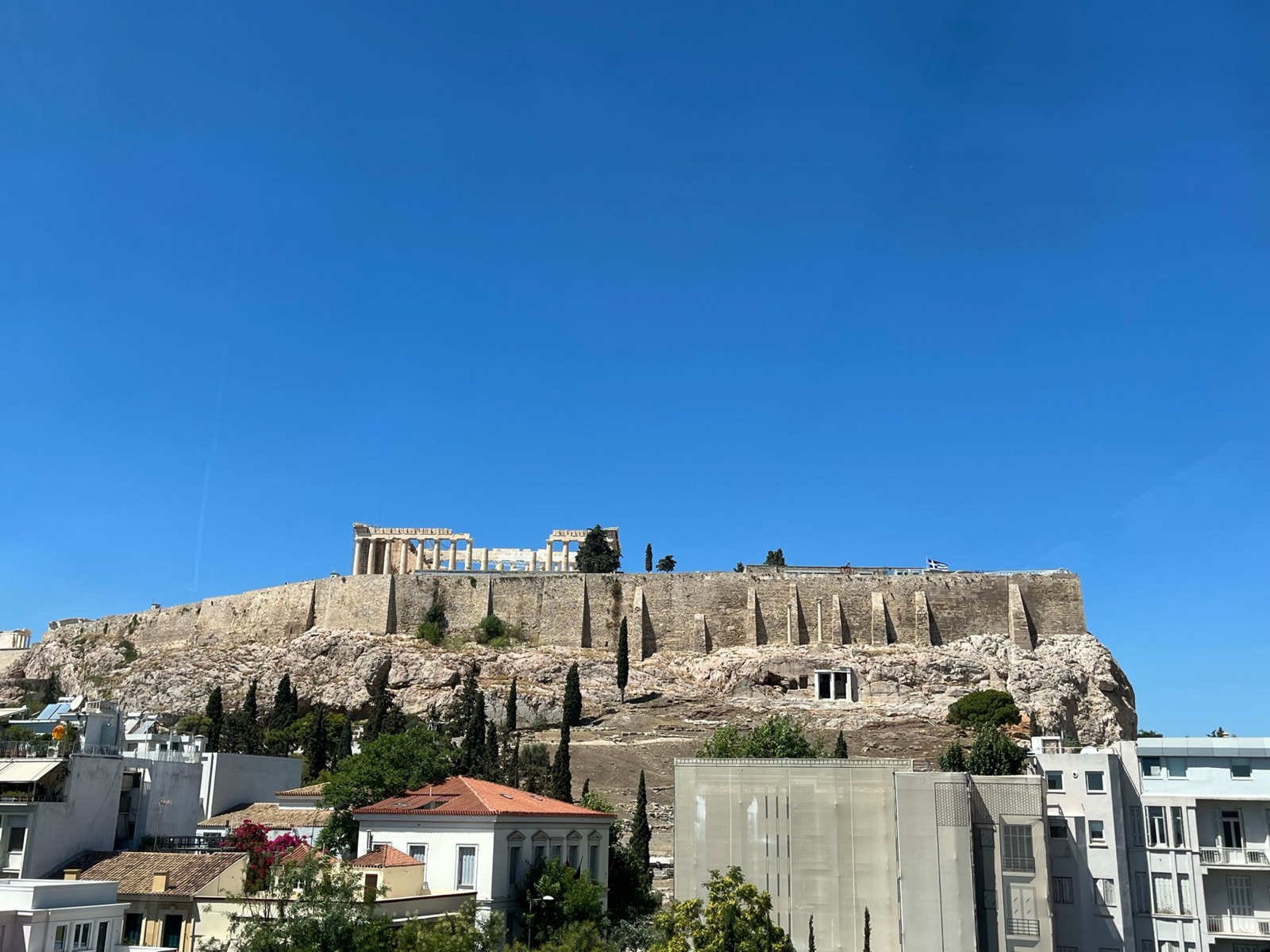 The Acropolis and Parthenon in daylight under a blue sky, seen from the Acropolis Museum, rooftops and trees in between
