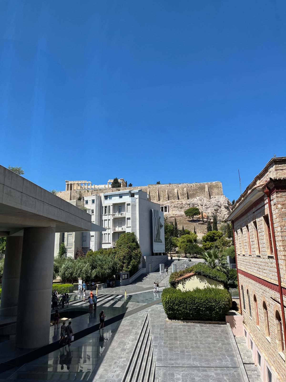 Ο διάδρομος εισόδου του Μουσείου Ακρόπολης με γυάλινο δάπεδο πάνω από αρχαίες ανασκαφές, ο Παρθενώνας ορατός στον βράχο από πάνω κάτω από γαλάζιο ουρανό