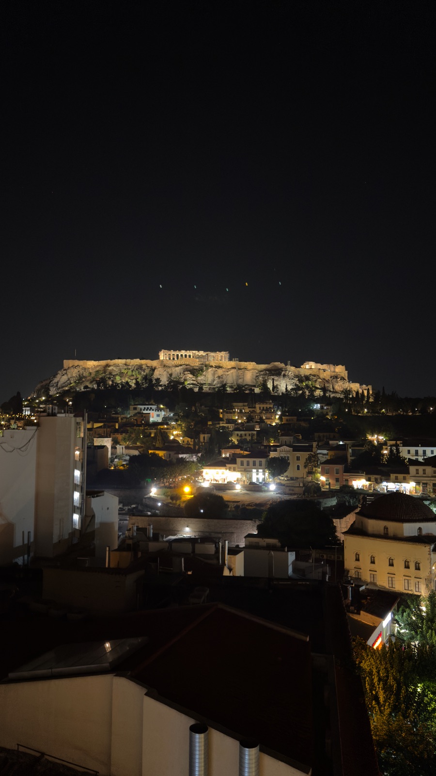 Η Ακρόπολη και ο Παρθενώνας φωτισμένοι τη νύχτα πάνω από τις στέγες της Πλάκας και του Μοναστηρακίου, ένας τρούλος τζαμιού κάτω δεξιά