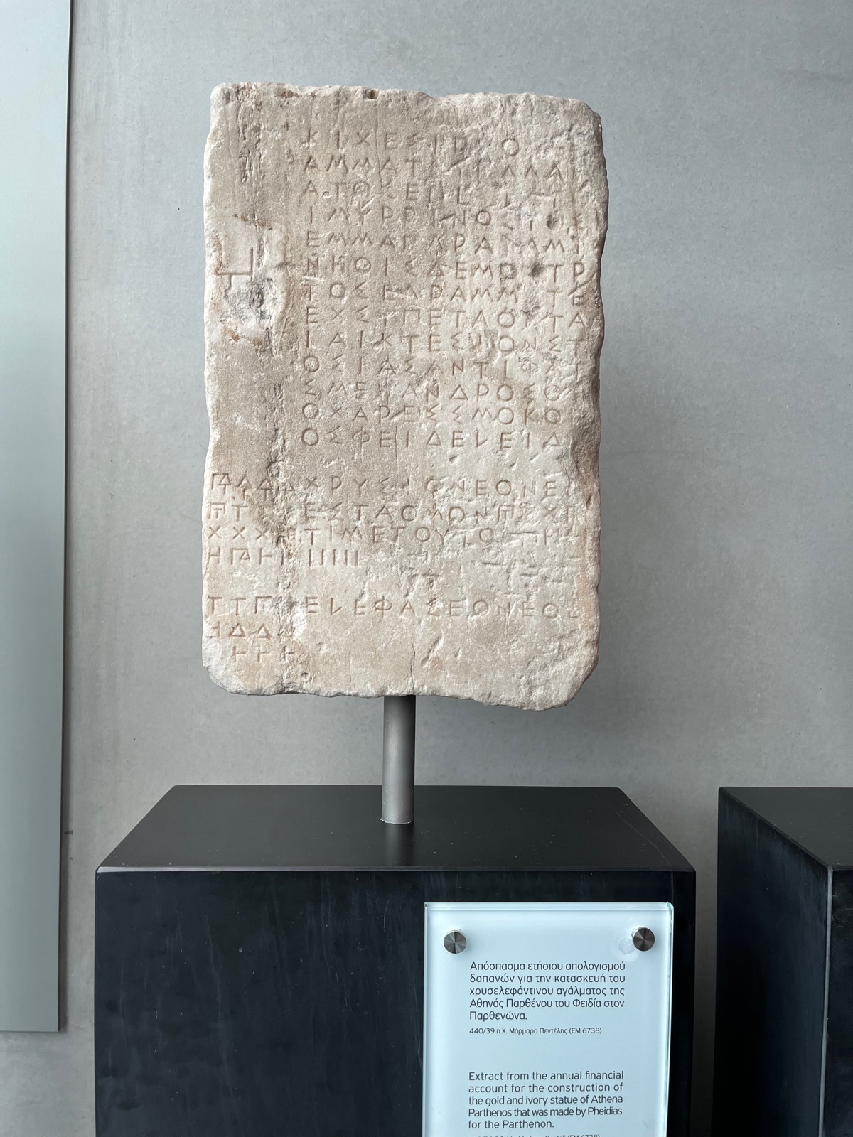 Μια ενεπίγραφη μαρμάρινη στήλη στο Μουσείο Ακρόπολης, η επιφάνειά της καλυμμένη με φθαρμένα αρχαία ελληνικά γράμματα, με επεξηγηματική πινακίδα από κάτω