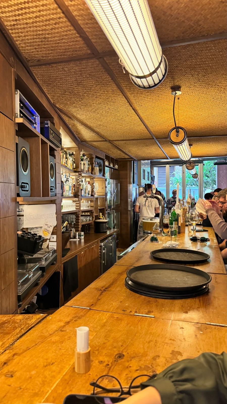 The interior of Birdman, a long wooden bar with turntables, shelves of glasses and spirits, and staff working under woven ceiling panels