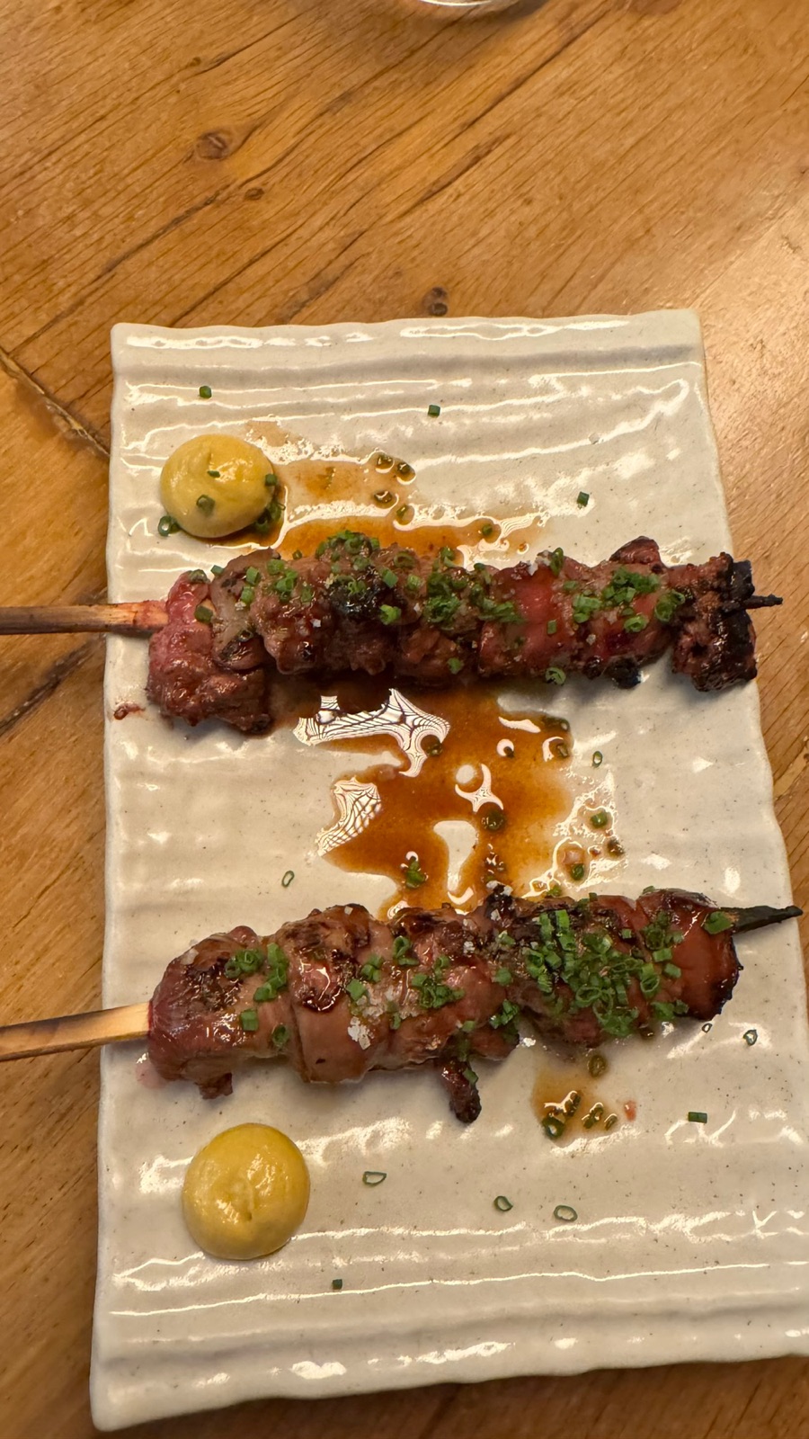Two glazed meat skewers scattered with chives on a ribbed ceramic plate, with two dots of mustard