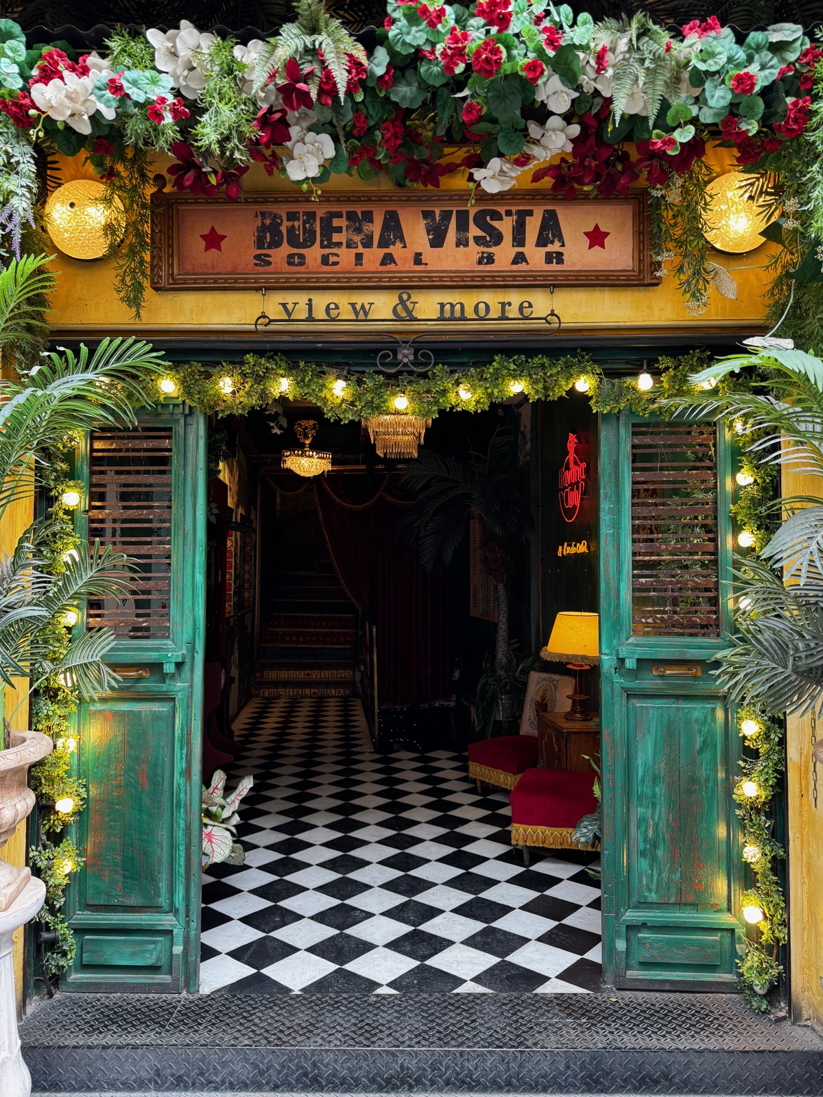 Η είσοδος του Buena Vista Social Bar με ζωγραφισμένη πινακίδα, πράσινες πόρτες-παντζούρια, γιρλάντες φώτων και τεχνητά λουλούδια γύρω από την πόρτα
