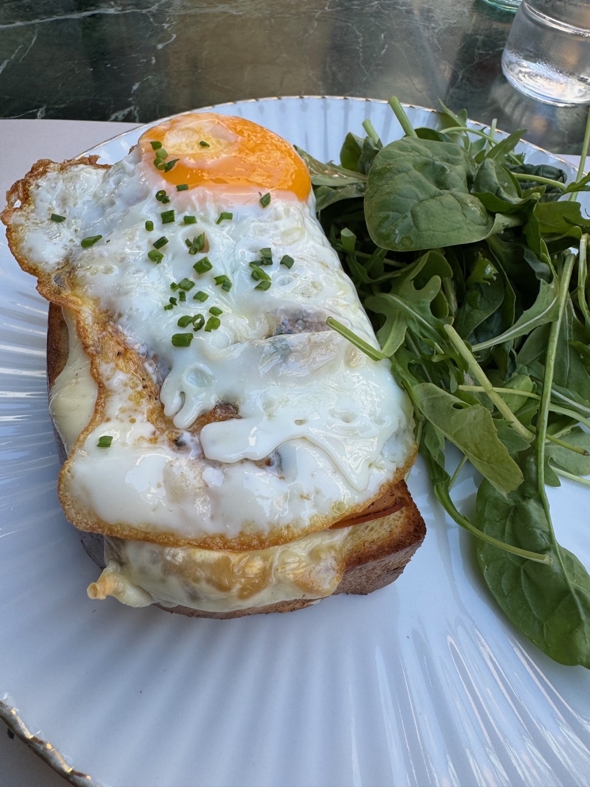 Ένα croque madame με τηγανητό αυγό από πάνω και λιωμένο τυρί στα πλάγια, με σαλάτα ρόκα και σπανάκι σε πιάτο με ραβδώσεις