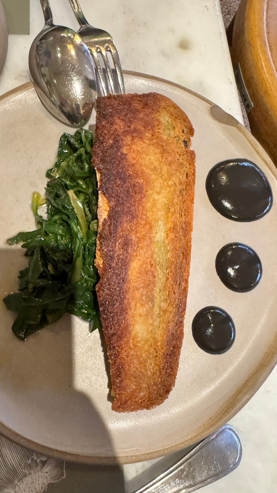 A golden fried starter over sauteed greens on a ceramic plate, with three round dots of black sauce lined up beside it