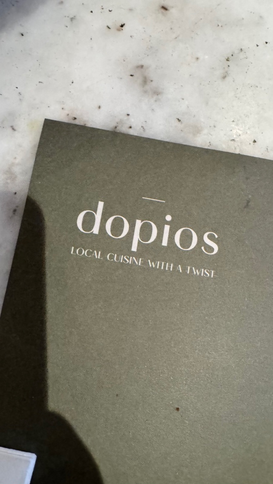 A dark green menu cover on a marble table reading dopios, local cuisine with a twist