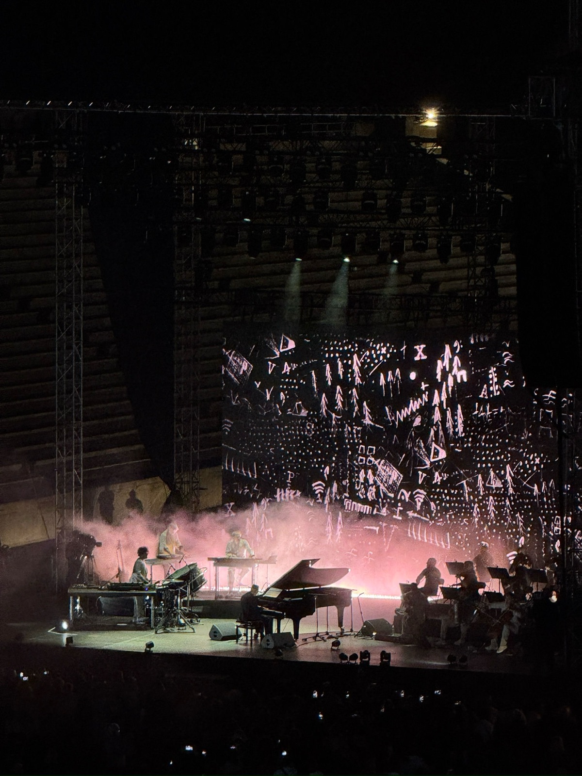 The same stage in pale pink light, a backdrop of white hand-drawn doodles behind the grand piano, keyboards and string players