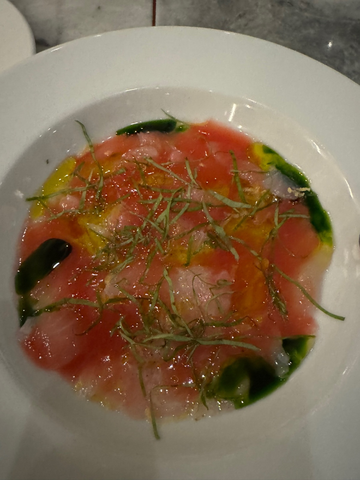 A bowl of pink fish crudo slices in a green herb oil, topped with shredded herbs