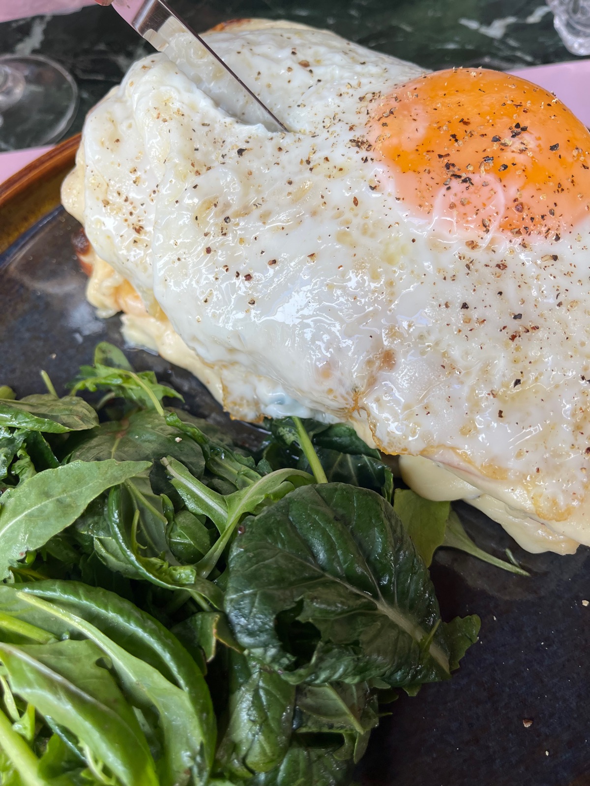 Ένα πιάτο brunch με δύο τηγανητά αυγά πάνω σε ψημένο σάντουιτς και μια συνοδεία πράσινης σαλάτας, σε σκούρο πιάτο πάνω σε μαρμάρινο τραπέζι