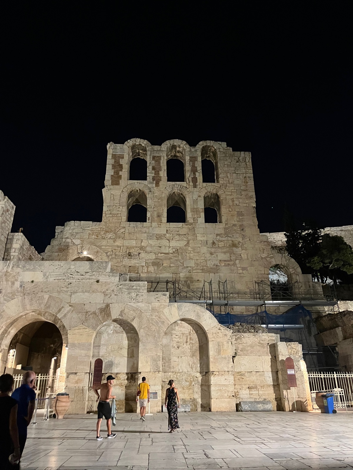 Η πέτρινη πρόσοψη του Ωδείου Ηρώδου του Αττικού φωτισμένη τη νύχτα, ο τοξωτός επάνω όροφός της να λάμπει πάνω στη σκοτεινή πλαγιά, λίγοι επισκέπτες στο μονοπάτι από κάτω