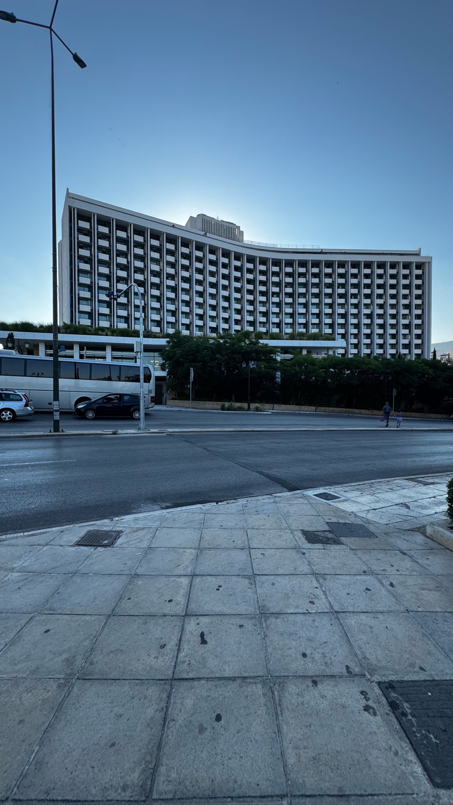 The curved modernist slab of the Hilton hotel seen from across an empty avenue in early morning light, the sun rising behind it