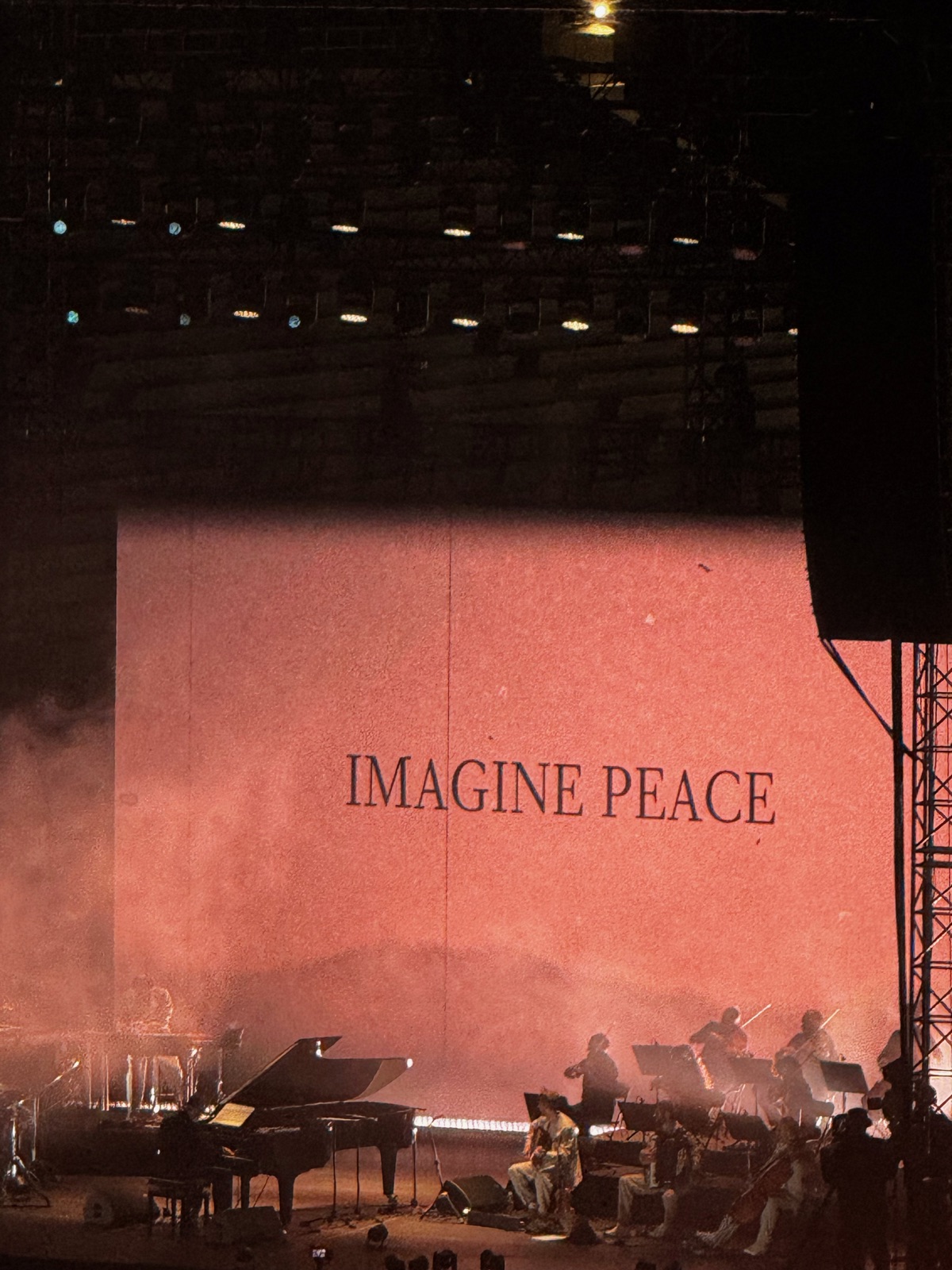 Μια σκηνή συναυλίας με τη φράση IMAGINE PEACE προβαλλόμενη σε ένα τεράστιο κόκκινα φωτισμένο φόντο, ένα πιάνο με ουρά αριστερά και έγχορδα σε σιλουέτα δεξιά
