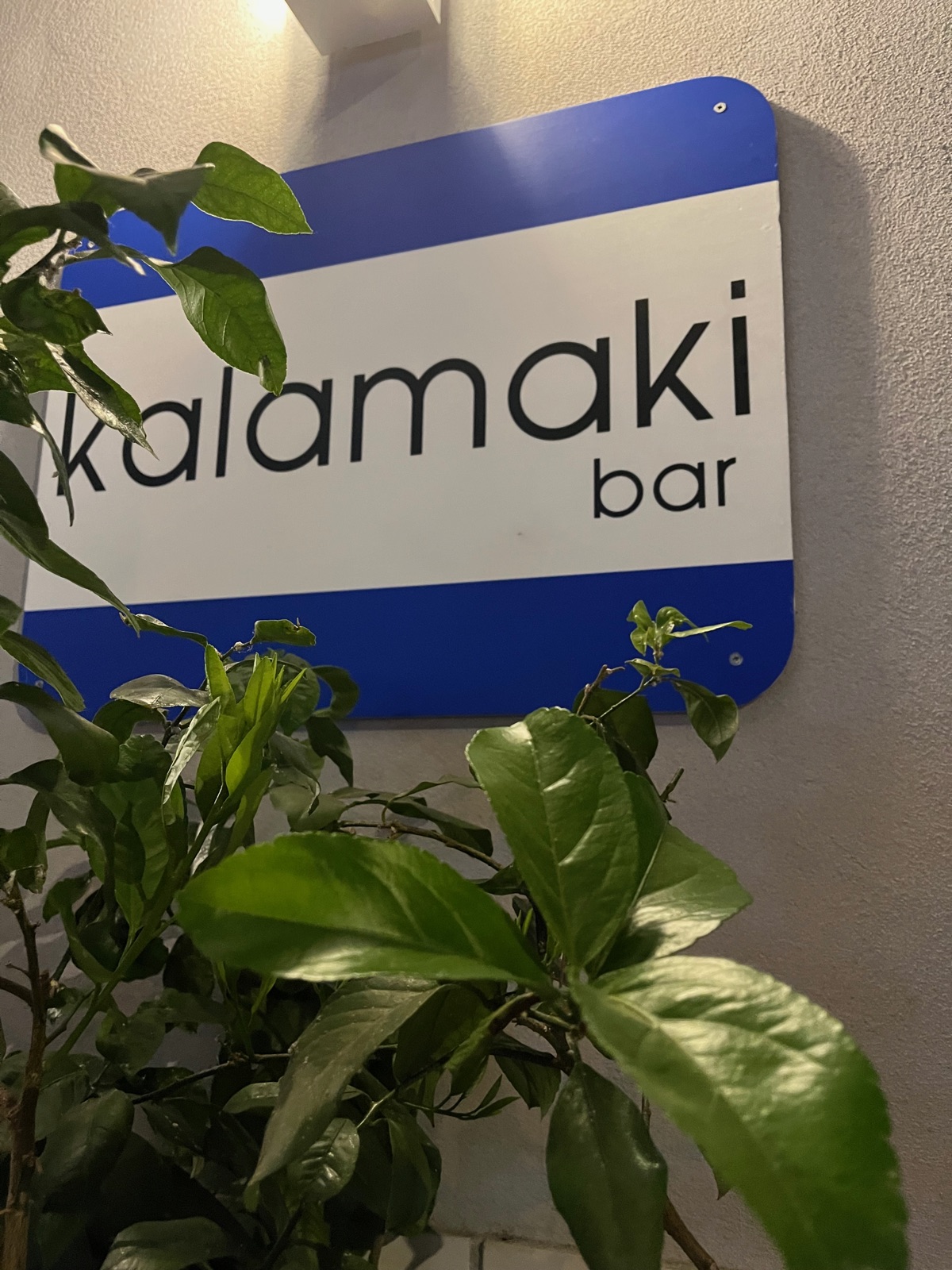 Μια μπλε και λευκή πινακίδα που γράφει kalamaki bar στερεωμένη σε τοίχο πίσω από τα κλαδιά μιας λεμονιάς