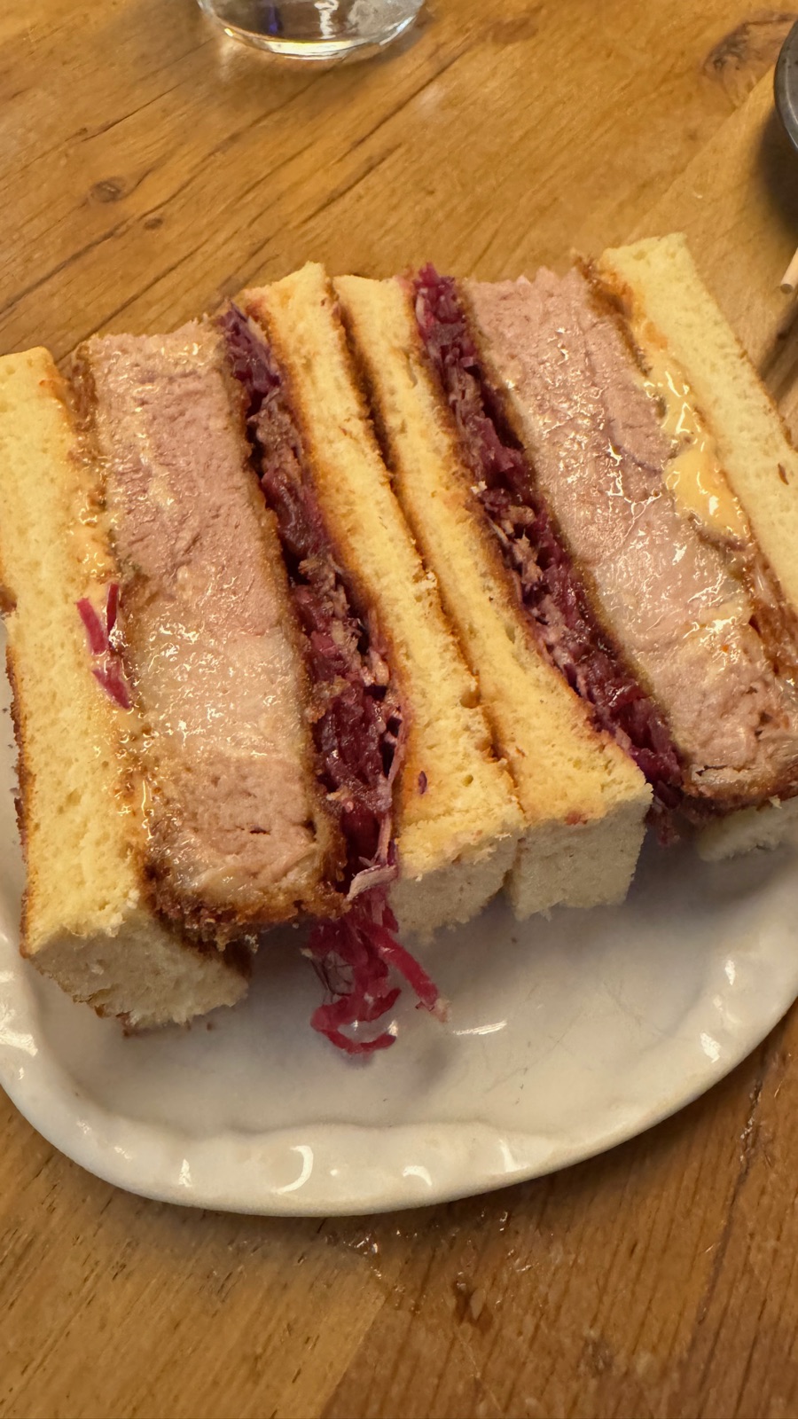 Ένα katsu sando κομμένο σε χοντρές φέτες σε λευκό πιάτο, πανέ χοιρινό και έντονα ροζ σαλάτα λάχανου πιεσμένα ανάμεσα σε χρυσό γαλακτερό ψωμί