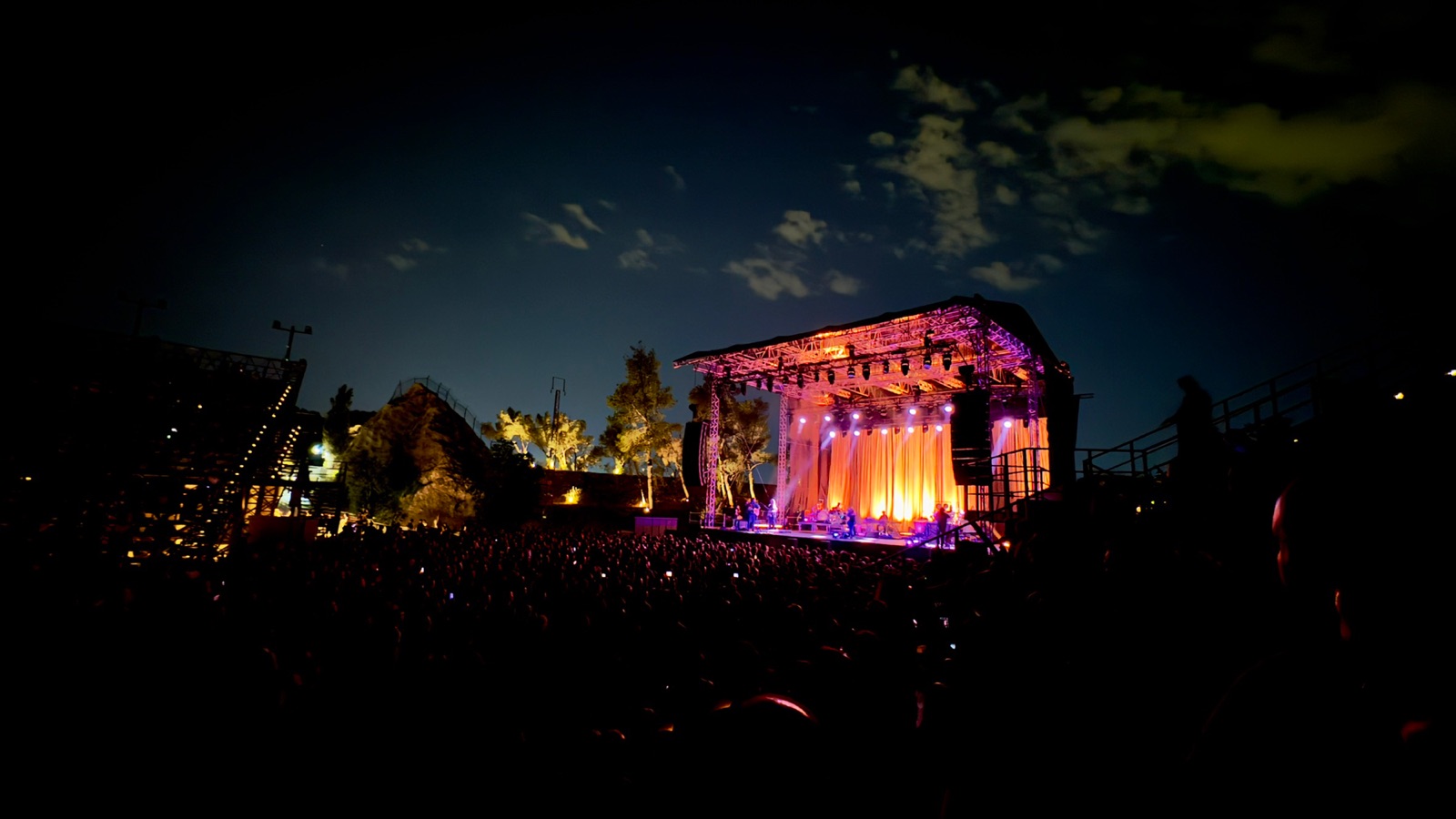 The Lycabettus stage at night under warm orange light, the dark silhouette of the quarry to the left and the crowd in shadow