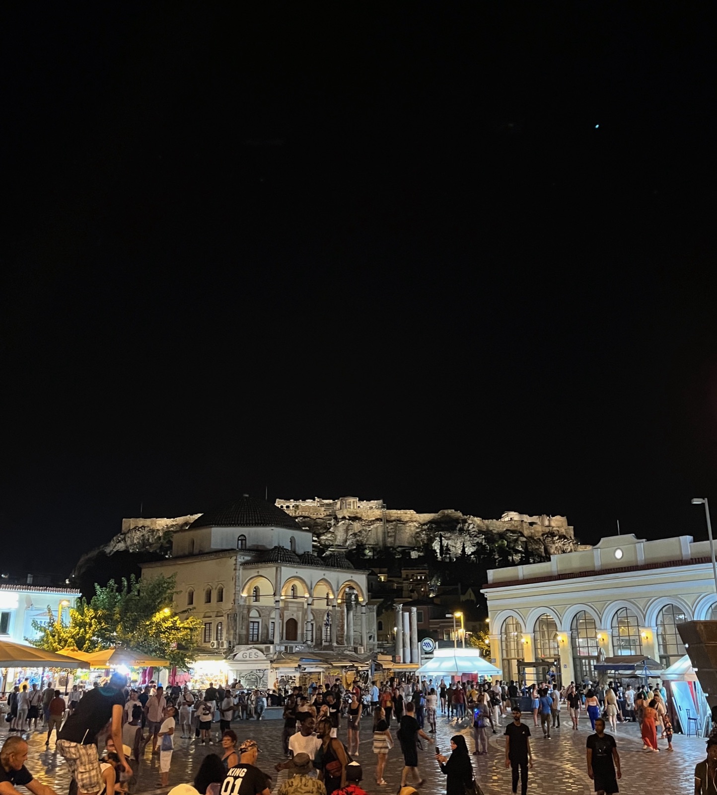 Η πλατεία Μοναστηρακίου τη νύχτα, το παλιό τζαμί και τα κτίρια της αγοράς φωτισμένα και η φωτισμένη Ακρόπολη από πάνω τους, αραιό πλήθος να διασχίζει την πλατεία