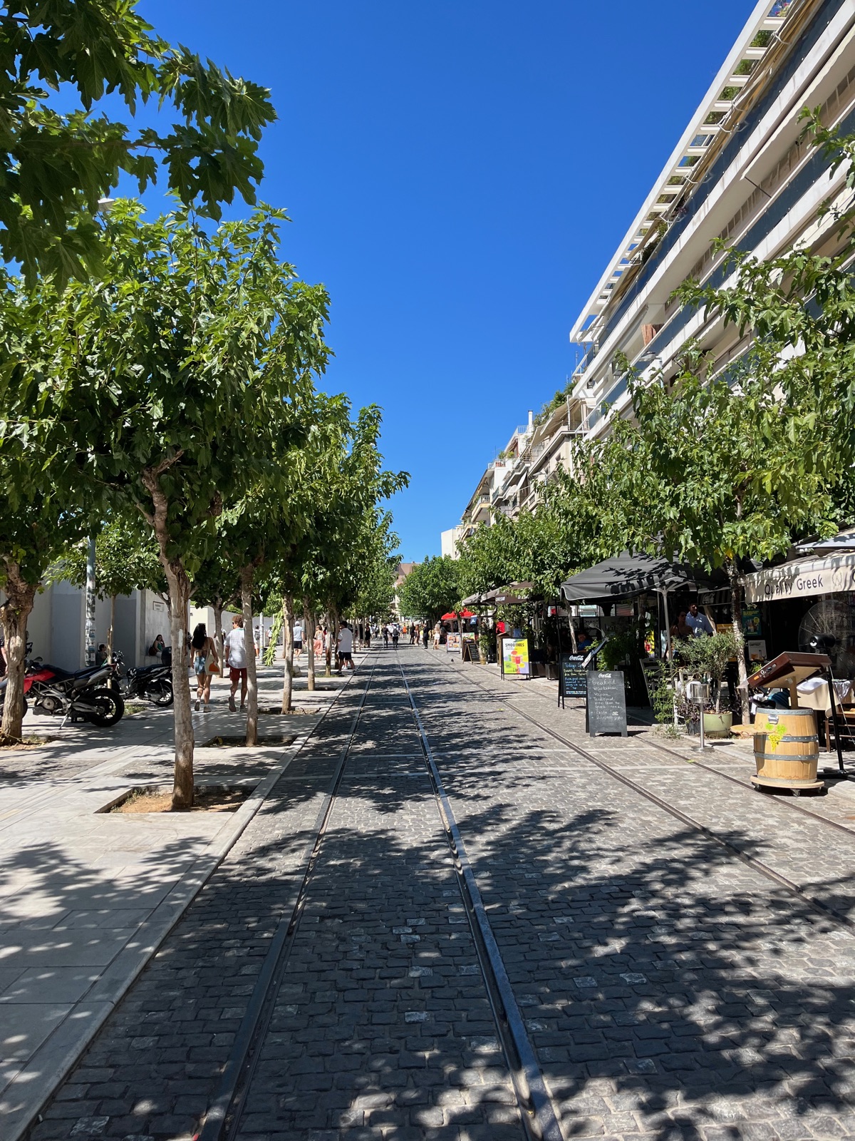 Ένας δεντροφυτεμένος πεζόδρομος κοντά στο Μουσείο Ακρόπολης με παλιές ράγες χωμένες στο πλακόστρωτο και καφέ στη δεξιά πλευρά