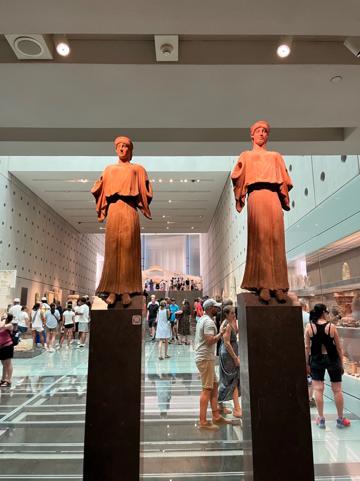 Two clay-colored statues on plinths inside the Acropolis Museum, visitors crossing a glass floor behind them