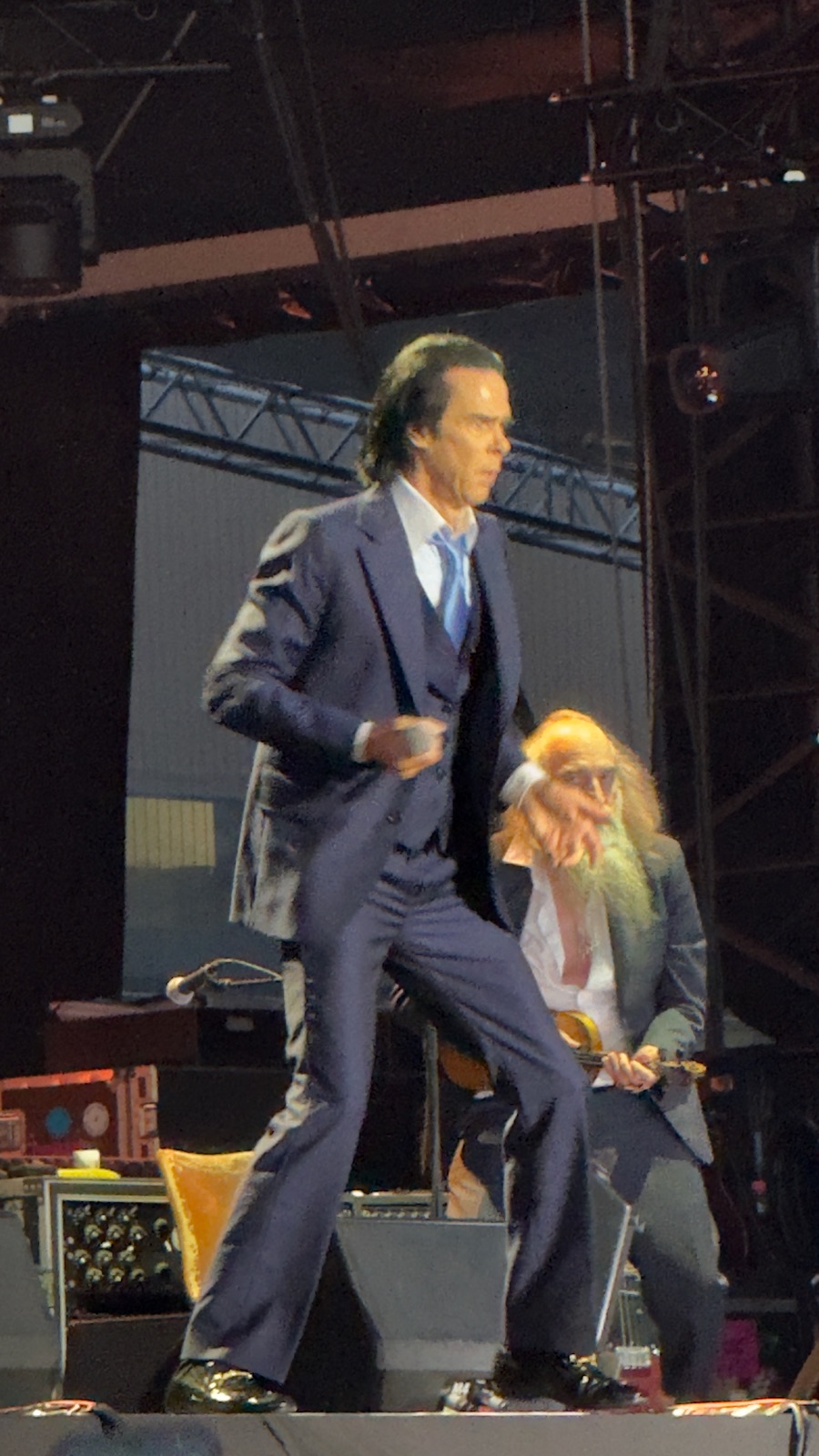 Ο Nick Cave με σκούρο τρίκουστο κοστούμι να διασχίζει τη σκηνή, ένα γενειοφόρο μέλος της μπάντας να παίζει πίσω του