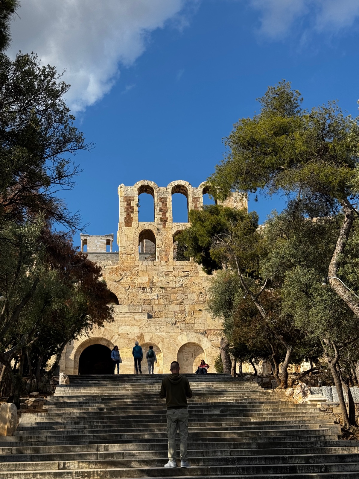 Η τοξωτή πέτρινη πρόσοψη του Ωδείου Ηρώδου του Αττικού στο απογευματινό φως, ιδωμένη από τα πλατιά σκαλιά από κάτω με λίγους ανθρώπους να ανεβαίνουν