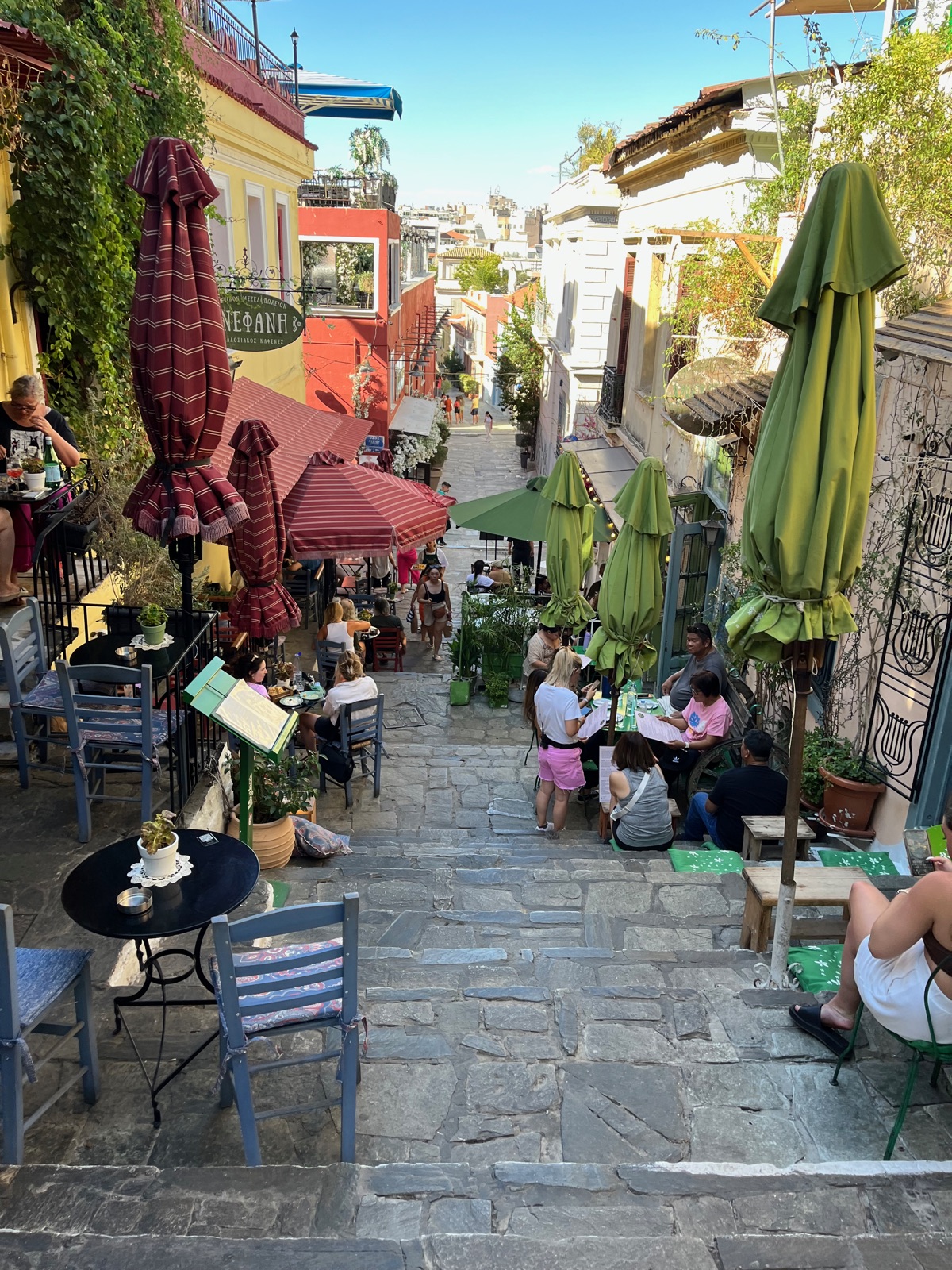 Ένα σκαλωτό σοκάκι στην Πλάκα γεμάτο τραπέζια ταβέρνας και ομπρέλες, πρασινάδα από πάνω και πινακίδες στην κορυφή της σκάλας
