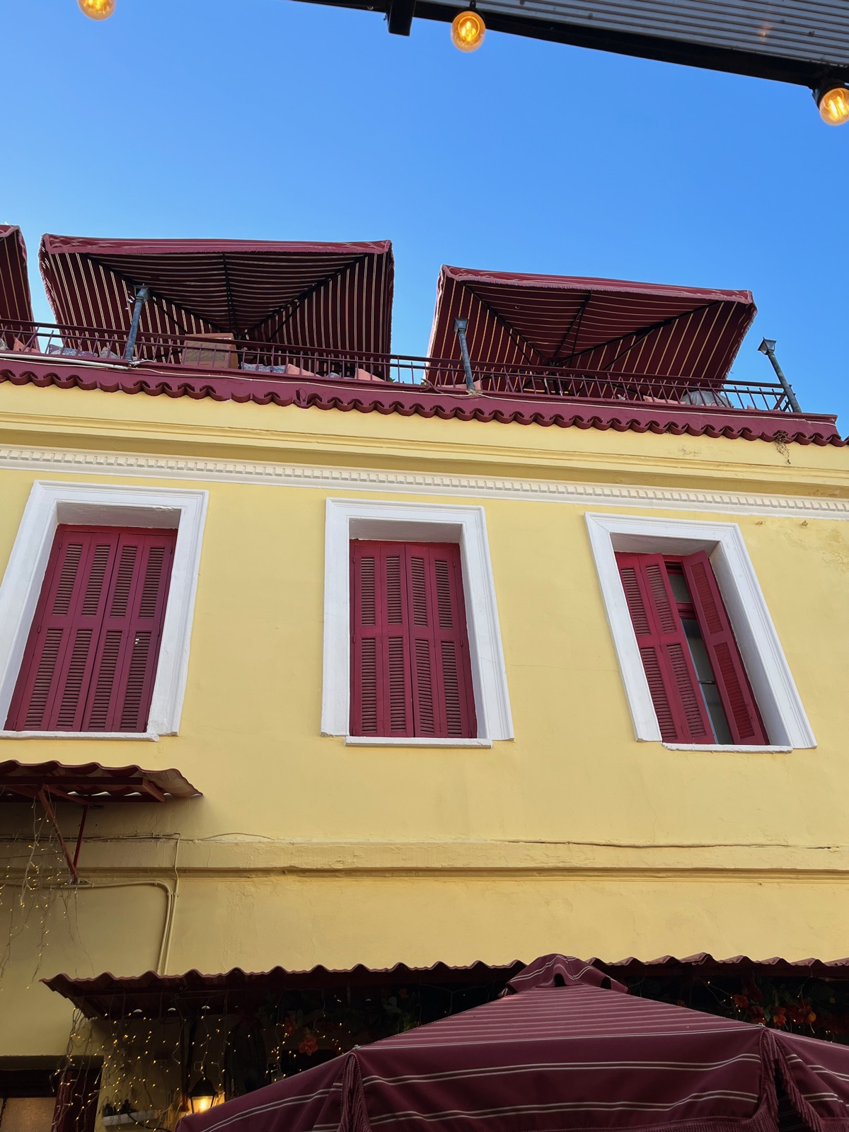 Ένα κίτρινο νεοκλασικό σπίτι στην Πλάκα με σκούρα κόκκινα παντζούρια, μια ταράτσα με ριγέ ομπρέλες και γιρλάντες φώτων από πάνω