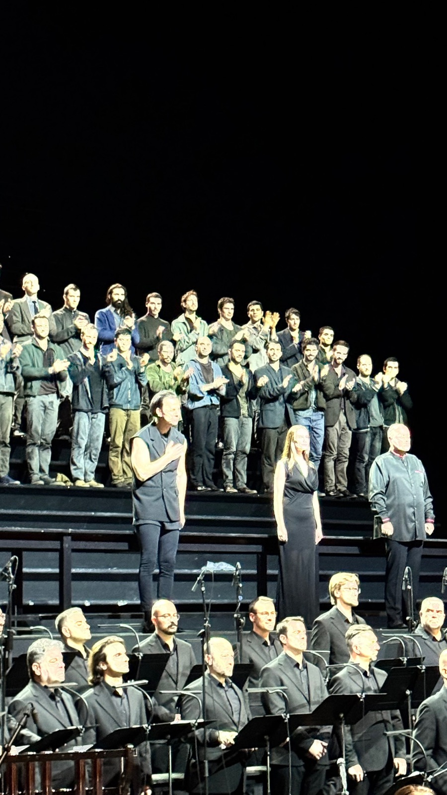 Three soloists taking their bow in front of the choir, a conductor in black with a hand on his chest and a soprano in a long dark dress