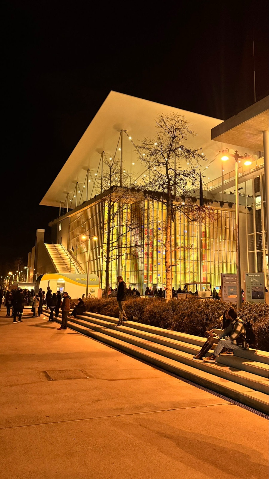 The Stavros Niarchos Cultural Center at night, glowing under its vast flat canopy, with broad steps, a food truck and people gathering outside