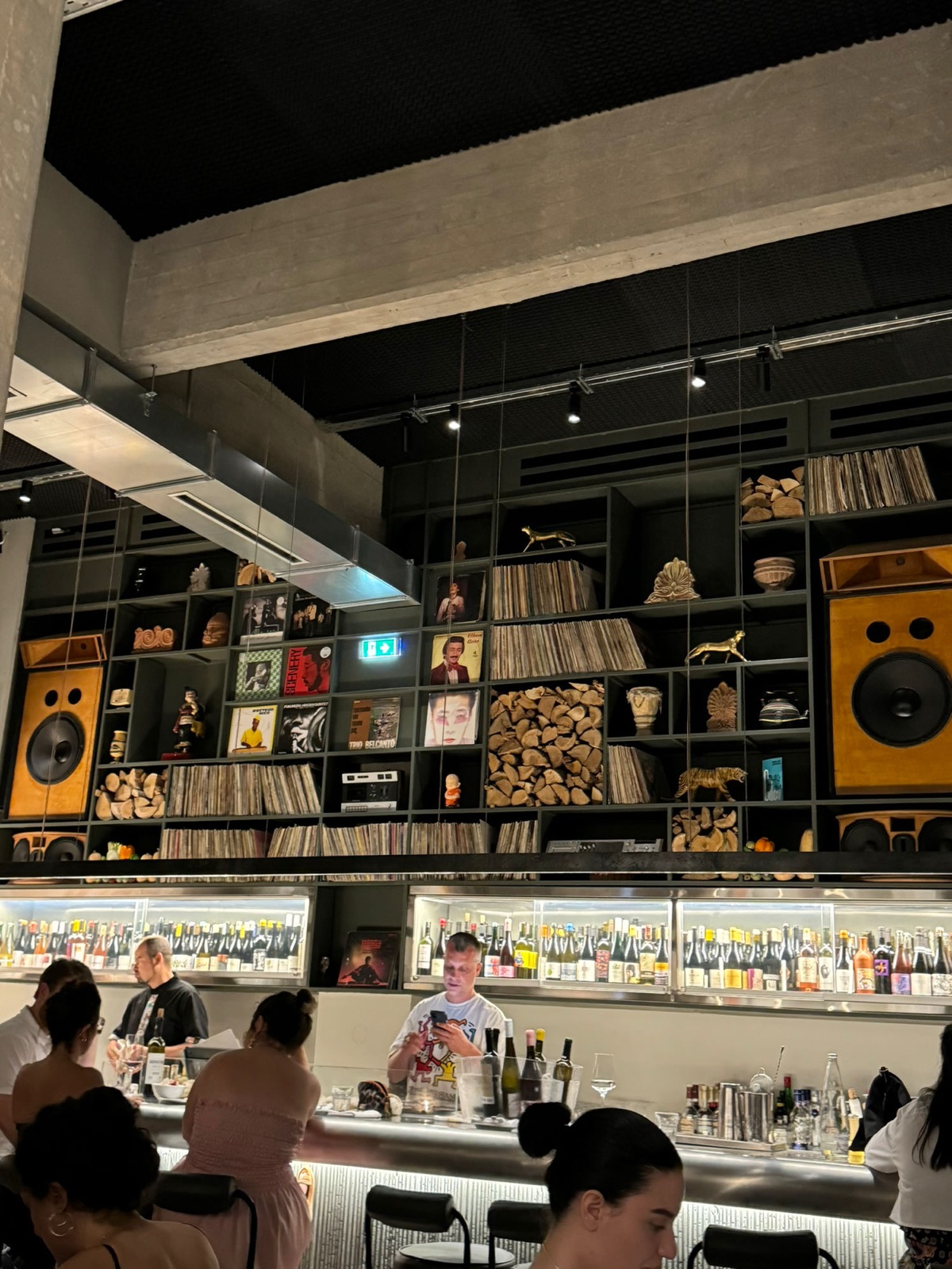 A bar interior with turntables, studio speakers and shelves of records and bottles behind a long wooden counter, staff working at the far end