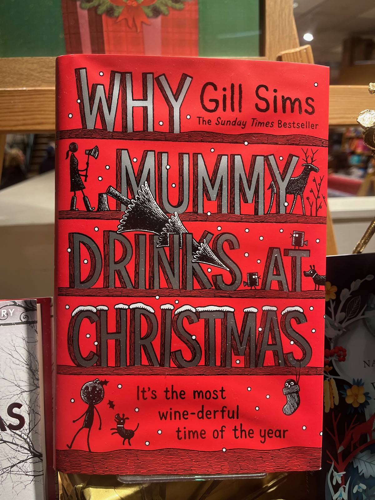 Ένα κόκκινο σκληρόδετο αντίτυπο του Why Mummy Drinks at Christmas της Gill Sims σε καβαλέτο σε βιβλιοπωλείο, με χριστουγεννιάτικα στολίδια γύρω του