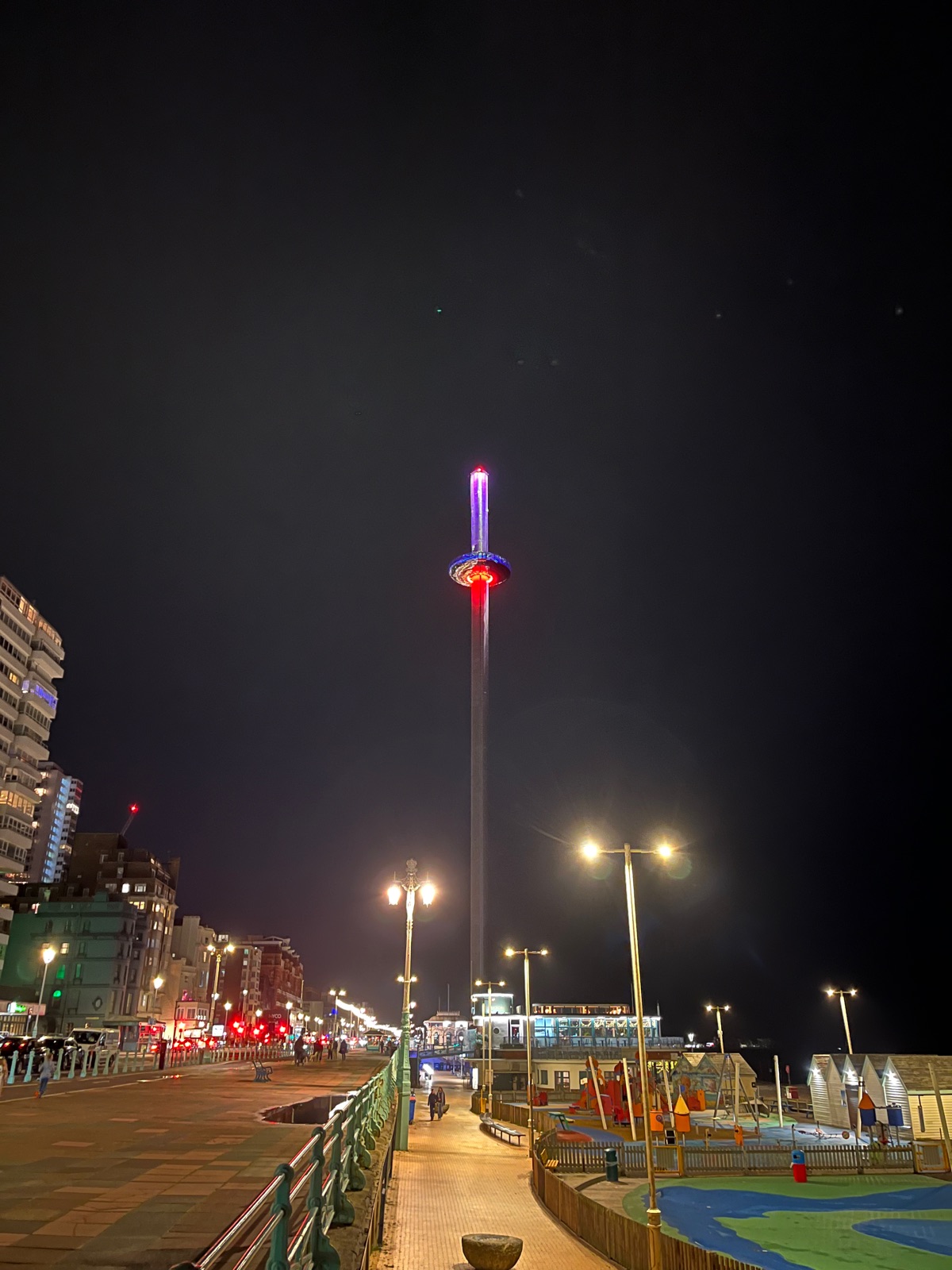 Ο πύργος i360 τη νύχτα φωτισμένος μωβ και κόκκινος σε όλο του το ύψος, πάνω από έναν φωτισμένο περίπατο και μια άδεια παιδική χαρά δίπλα στην παραλία