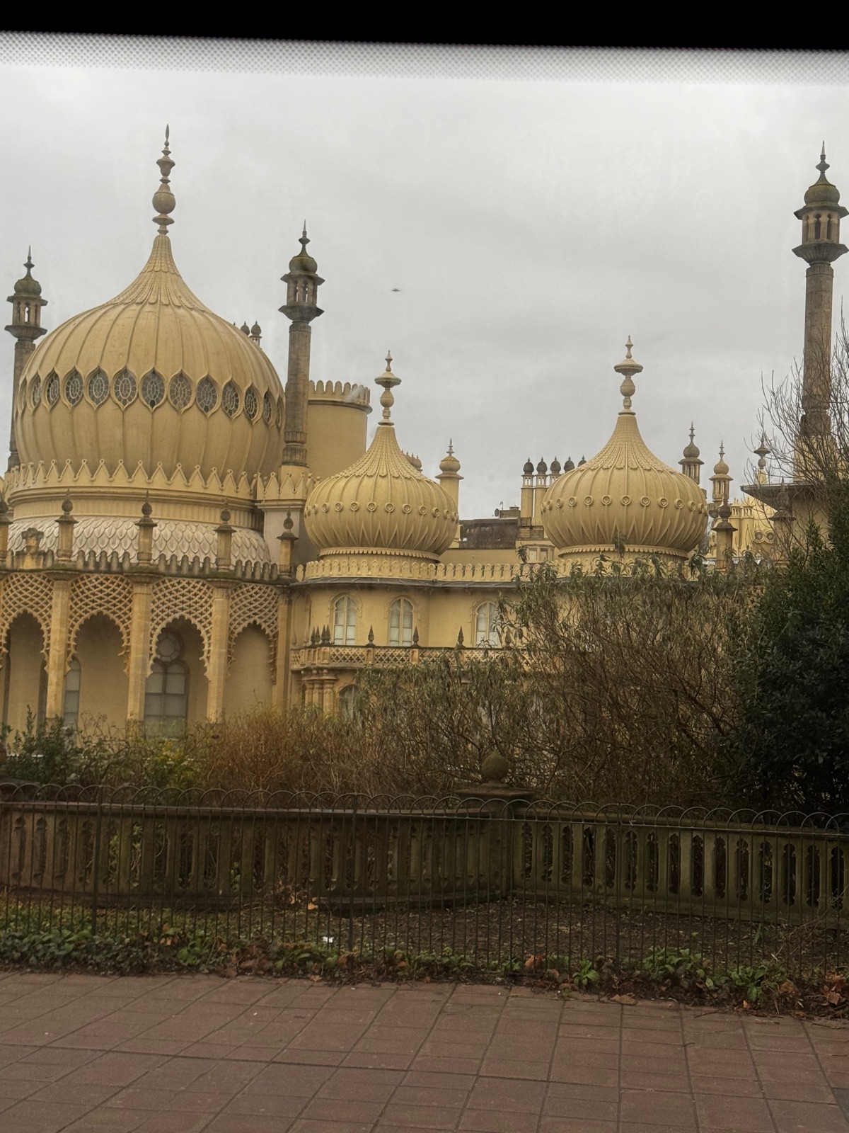 Οι χλωμοί θόλοι και μιναρέδες του Royal Pavilion να υψώνονται πάνω από θάμνους κήπου και σιδερένια κάγκελα κάτω από επίπεδο γκρίζο ουρανό, φωτογραφημένοι μέσα από παράθυρο με σκούρα σκιά κουφώματος στην κορυφή