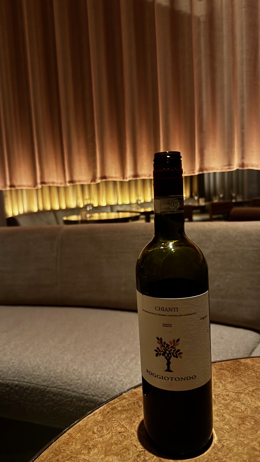 Ένα μπουκάλι Poggiotondo Chianti 2023 σε μικρό στρογγυλό τραπέζι σε αμυδρά φωτισμένο εστιατόριο, ένας καμπύλος καναπές και μια ροζ κουρτίνα να λάμπουν πίσω του