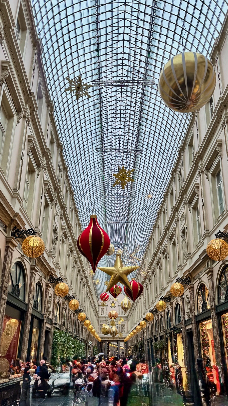 Η γυάλινη καμαρωτή στέγη των Galeries Royales Saint-Hubert με μια γιγάντια χρυσή μπάλα και συστάδες από κόκκινα και χρυσά στολίδια και αστέρια κρεμασμένα κατά μήκος της στοάς, ψωνιστές θολοί από κάτω