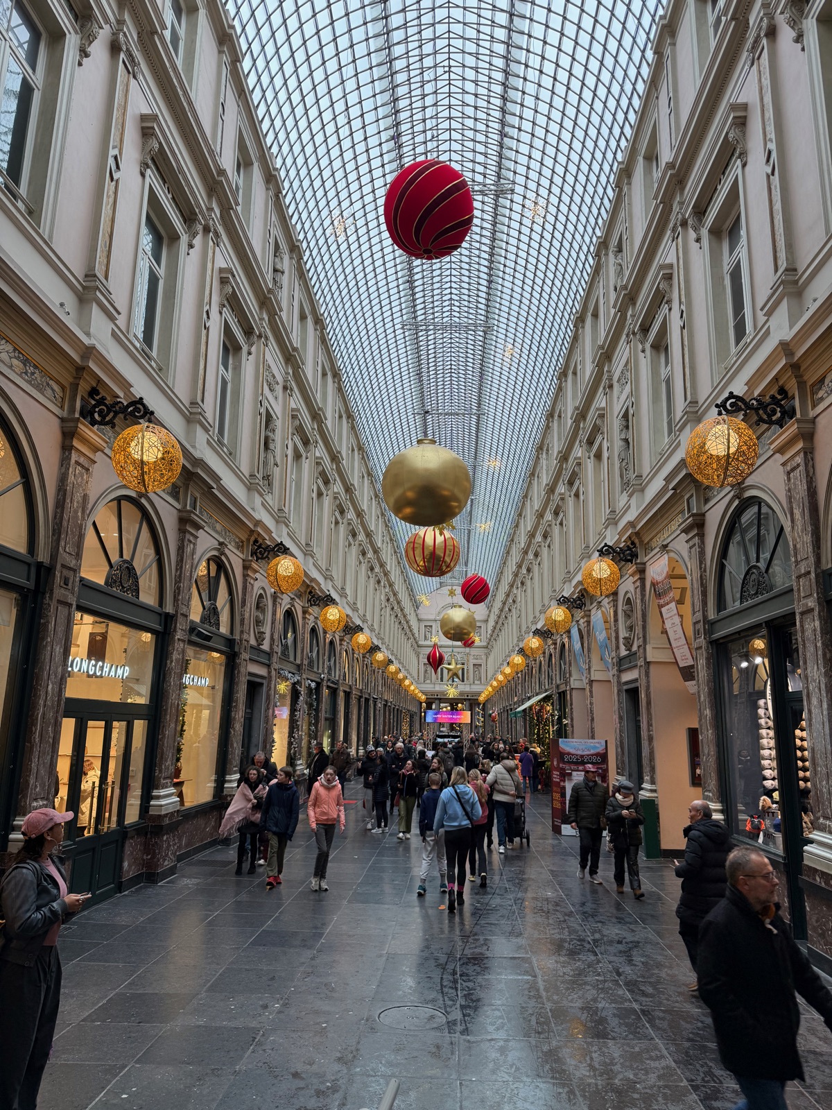 Η μακρινή θέα κατά μήκος των Galeries Royales Saint-Hubert, γυάλινη στέγη από πάνω, χρυσά στολίδια να χάνονται στο βάθος πάνω από ψωνιστές και φωτισμένες βιτρίνες