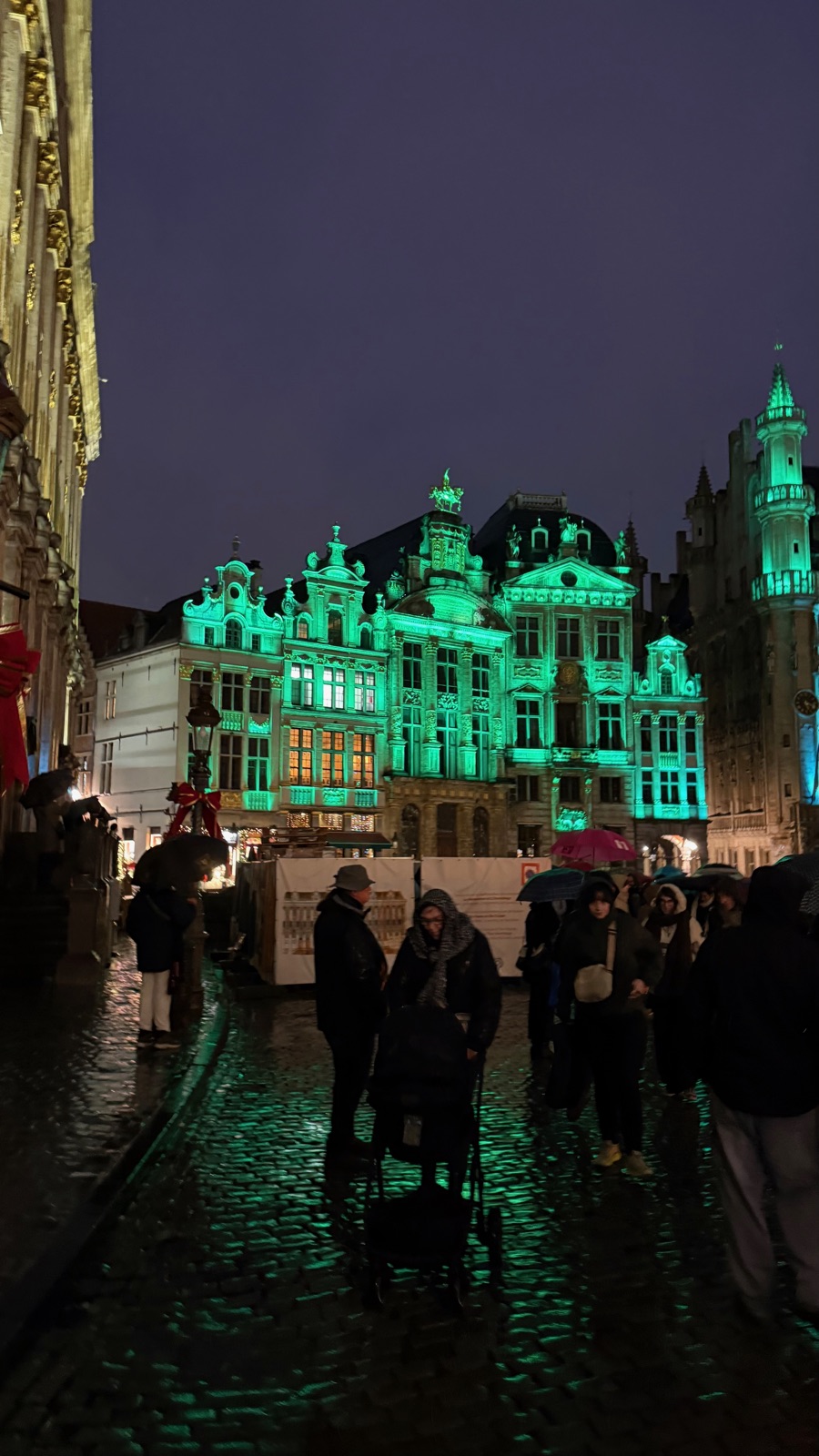 Σπίτια συντεχνιών στην Grand Place λουσμένα σε πράσινο φως τη νύχτα, ένα επιχρυσωμένο έφιππο άγαλμα να λάμπει στο κεντρικό αέτωμα, κόσμος με ομπρέλες στα βρεγμένα λιθόστρωτα από κάτω