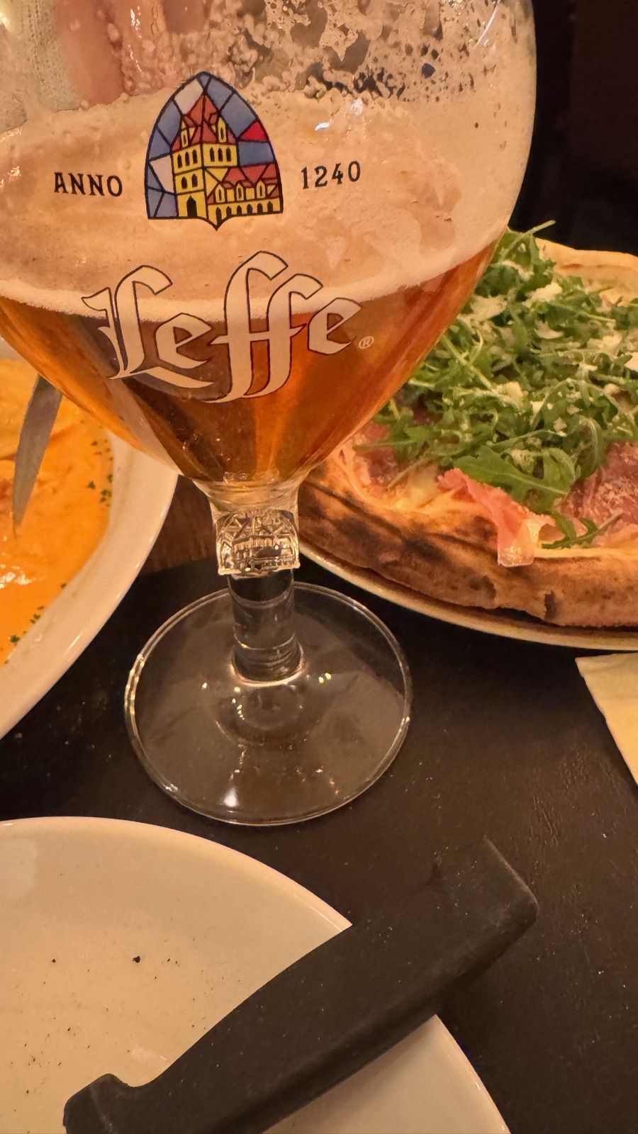 Ένα κύπελλο μπίρας Leffe με το λογότυπο του αβαείου Anno 1240, μπροστά από μια λεπτή τάρτα με ρόκα, ζαμπόν και ξυσμένο τυρί σε σκούρο τραπέζι