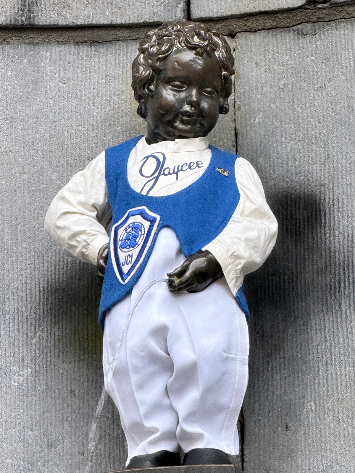 Κοντινή λήψη του Manneken Pis με λευκό πουκάμισο με καλλιγραφικά γράμματα Jaycee και βασιλικό μπλε γιλέκο με έμβλημα ασπίδας JCI, νερό να βγαίνει τοξωτά από το σιντριβάνι