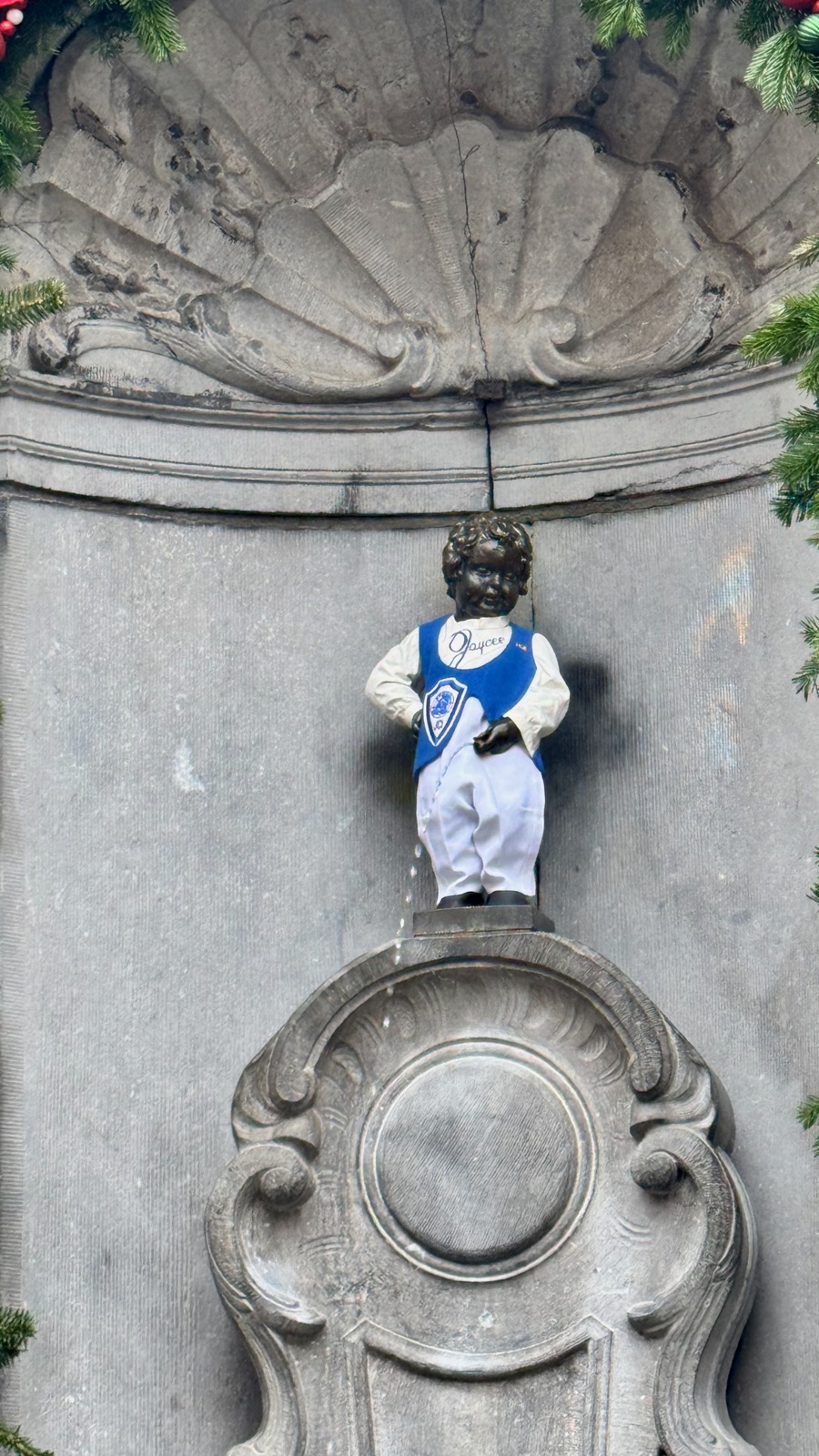 Το σιντριβάνι Manneken Pis στη σκαλισμένη πέτρινη κόγχη του στολισμένη με γιρλάντα από αειθαλή και κόκκινες μπάλες, το μικρό άγαλμα ντυμένο με μπλε και λευκή στολή