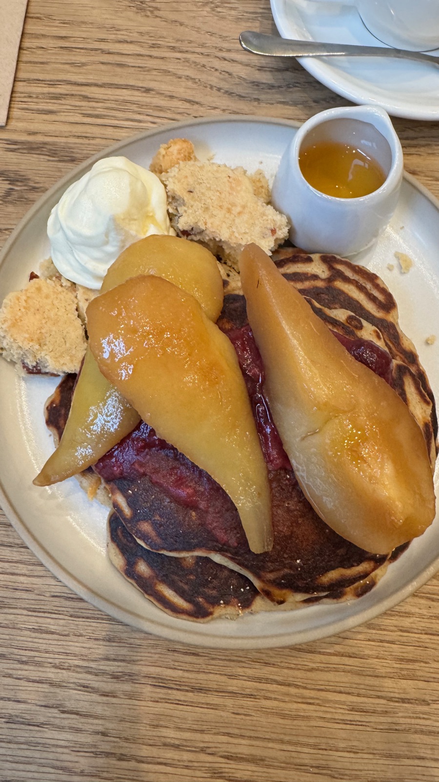 Μια στοίβα από pancakes με γλασαρισμένα μισά αχλάδια ποσέ, σκούρα κομπόστα φρούτων, σαντιγί και κράμπλ, με ένα μικρό κανάτι σιρόπι στο πιάτο