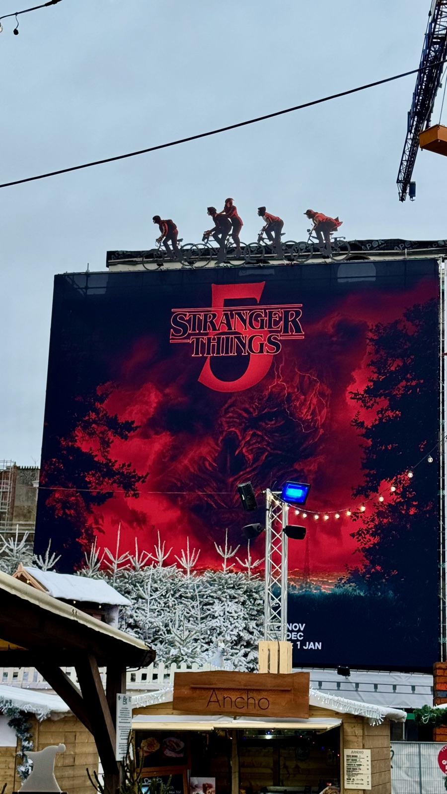 Μια γιγάντια σκοτεινή αφίσα του Stranger Things με σιλουέτες παιδιών σε ποδήλατα κατά μήκος της πάνω άκρης της, ένα σπιτάκι χριστουγεννιάτικης αγοράς και χιονισμένα τεχνητά έλατα από κάτω, ένας γερανός από πάνω