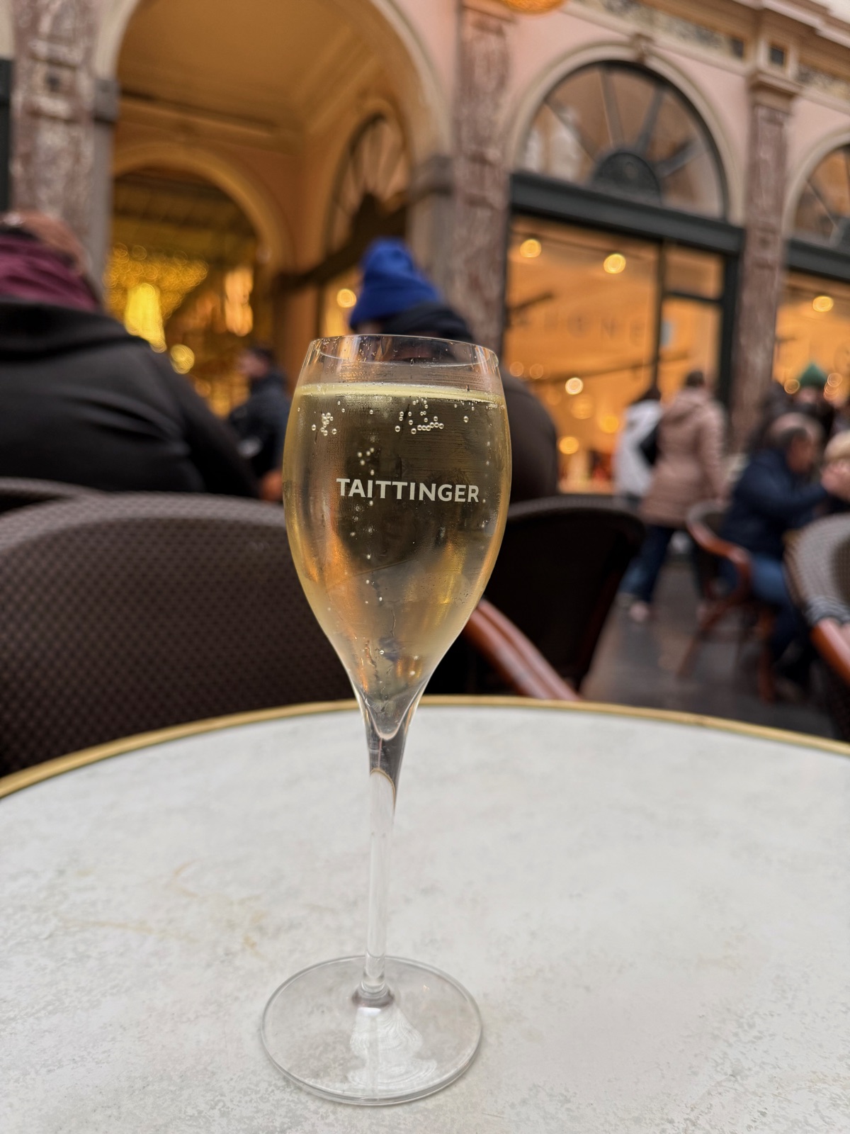 Ένα ποτήρι σαμπάνιας με την ένδειξη Taittinger, πάνω σε μαρμάρινο τραπεζάκι μπιστρό στη στοά, το πλήθος του καφέ και οι φωτισμένες βιτρίνες θολά πίσω
