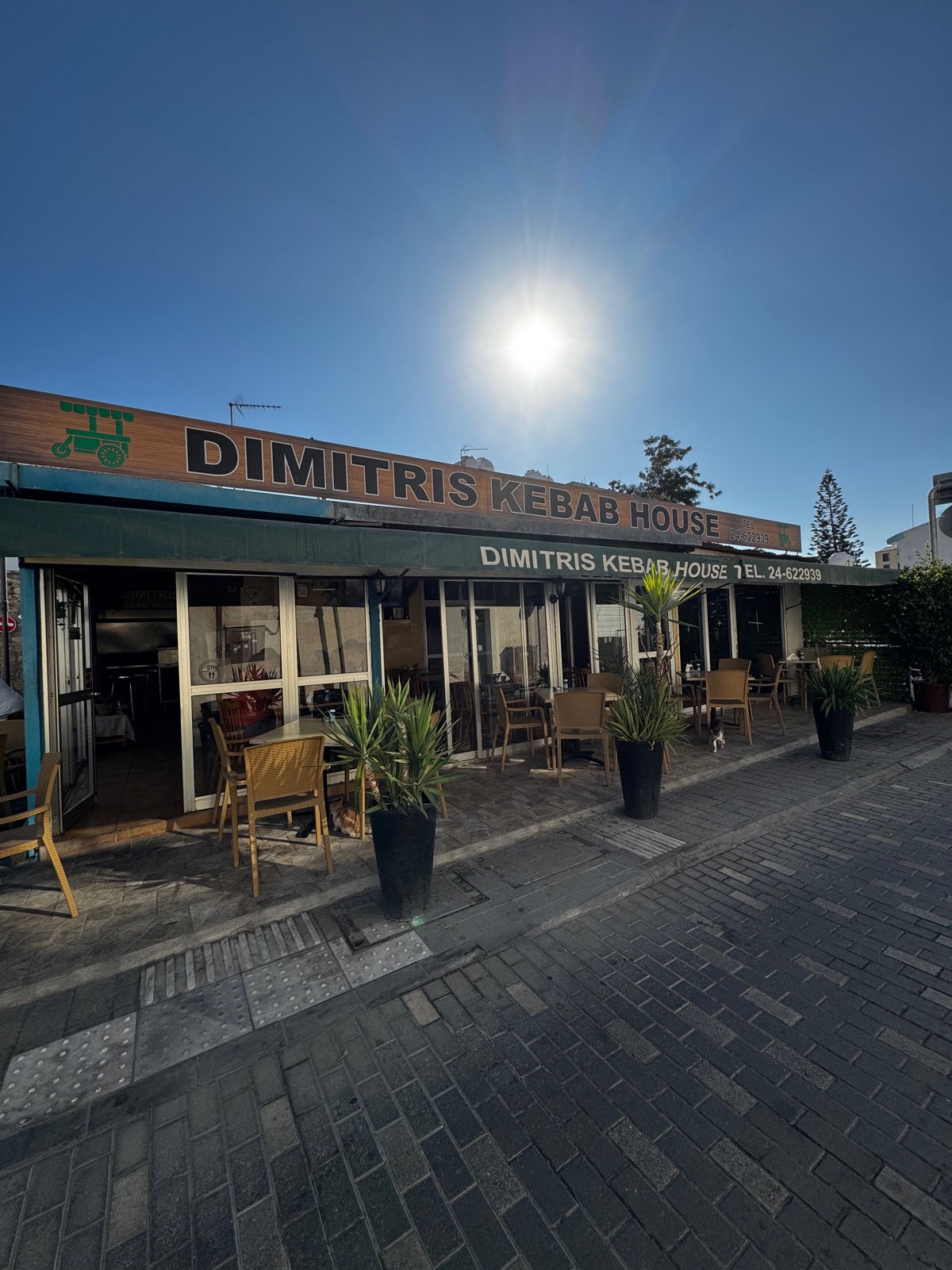 Η γωνιακή πρόσοψη του Dimitris Kebab House στη Λάρνακα με πράσινη τέντα, εξωτερικά τραπέζια, γλάστρες και μια γάτα δίπλα στις καρέκλες