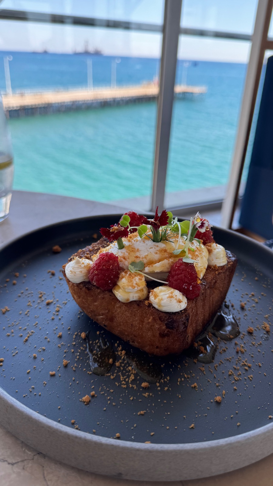 Ένα πιάτο french toast από μπριός με σαντιγί, βατόμουρα και λουλούδια από πάνω, σε τραπέζι δίπλα σε παράθυρο με θέα στη θάλασσα και μια ξύλινη προβλήτα