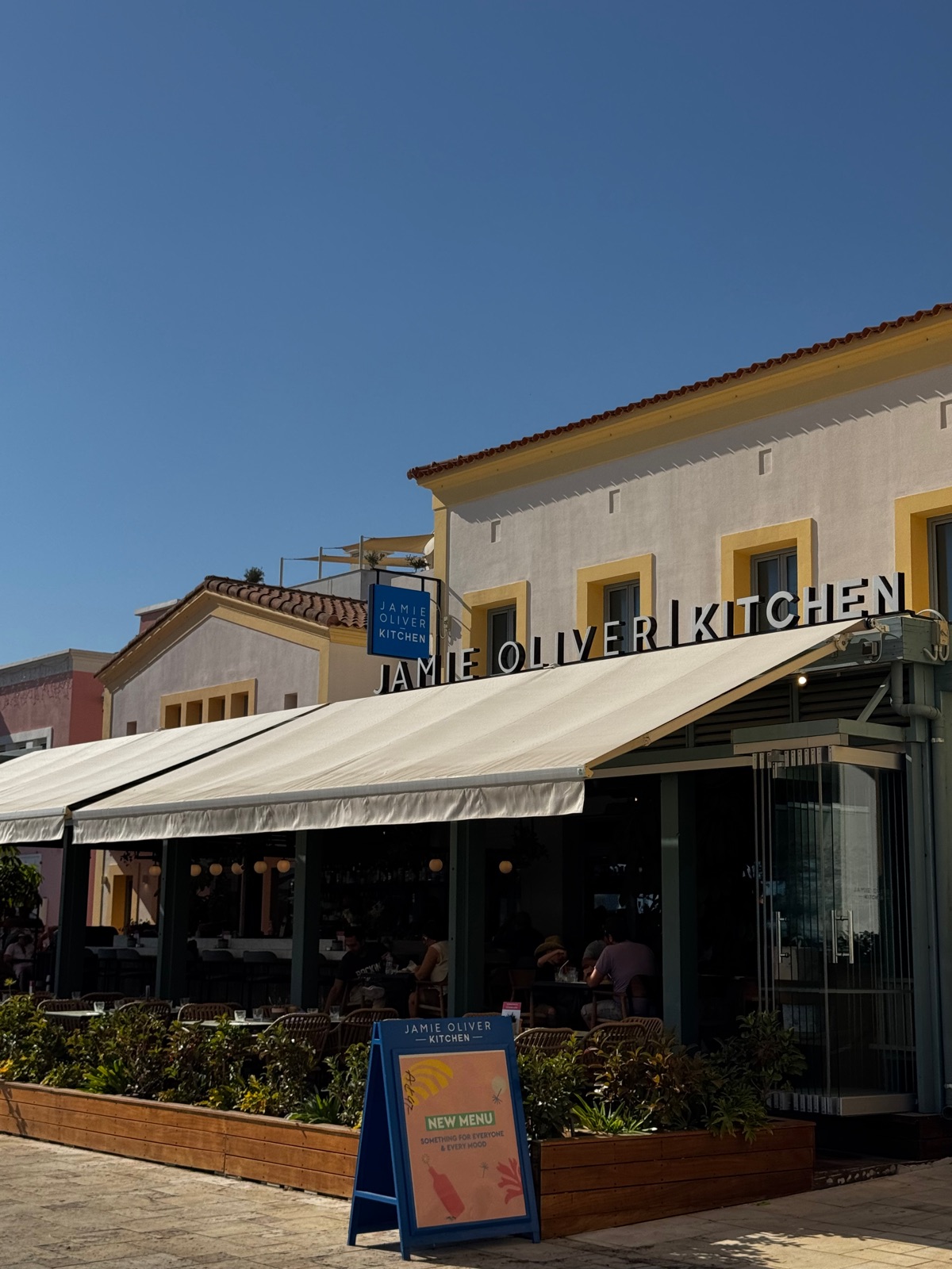 Η πρόσοψη ενός εστιατορίου Jamie Oliver Kitchen με λευκή τέντα και μια μπλε πινακίδα, κάτω από καθαρό ουρανό στη Λεμεσό