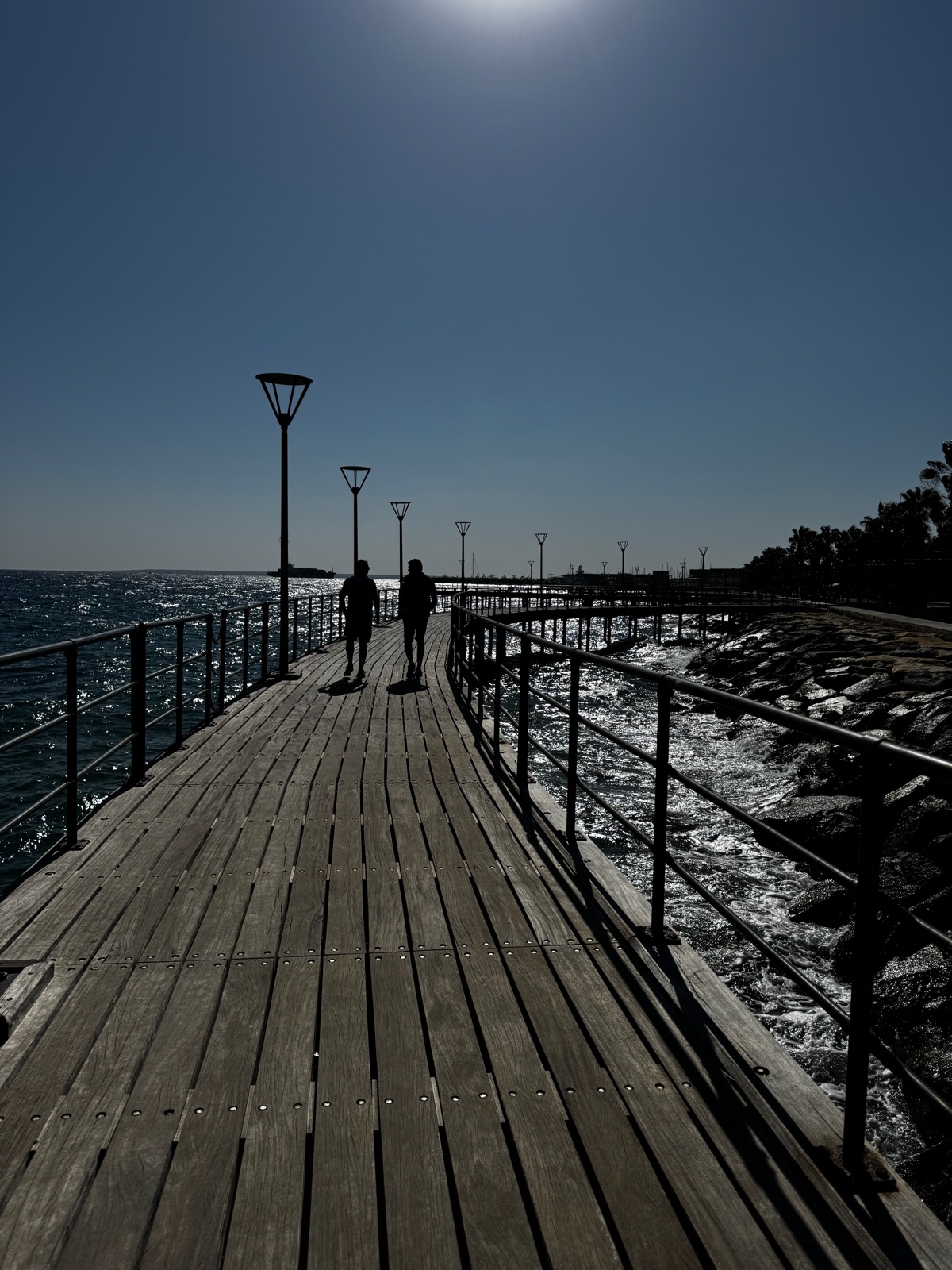 Μια χαμηλή ξύλινη προβλήτα να απλώνεται σε ασημένια θάλασσα κόντρα στον ήλιο, δύο μακρινές φιγούρες σε σιλουέτα στην άκρη της