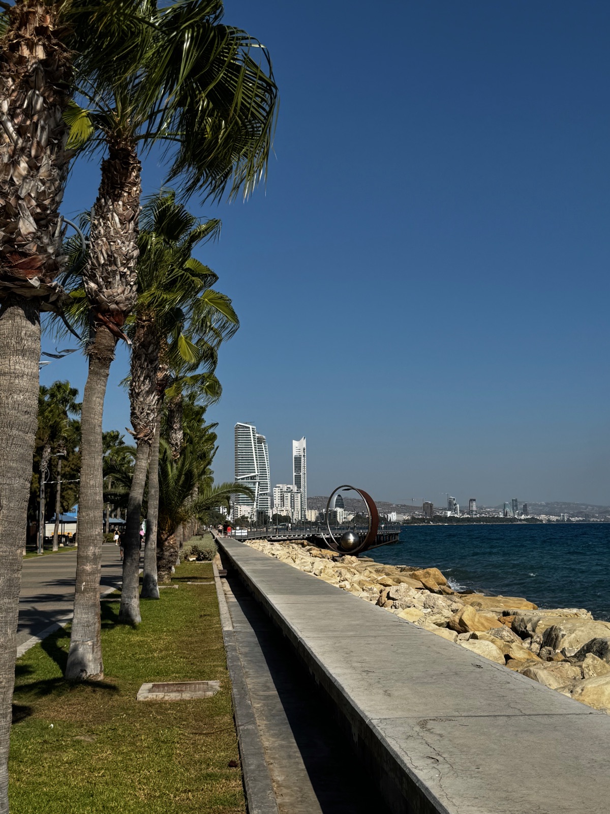 Ο παραλιακός περίπατος της Λεμεσού κατά μήκος ενός βραχώδους κυματοθραύστη, φοίνικες και ένα φανάρι, ψηλοί πύργοι στο βάθος