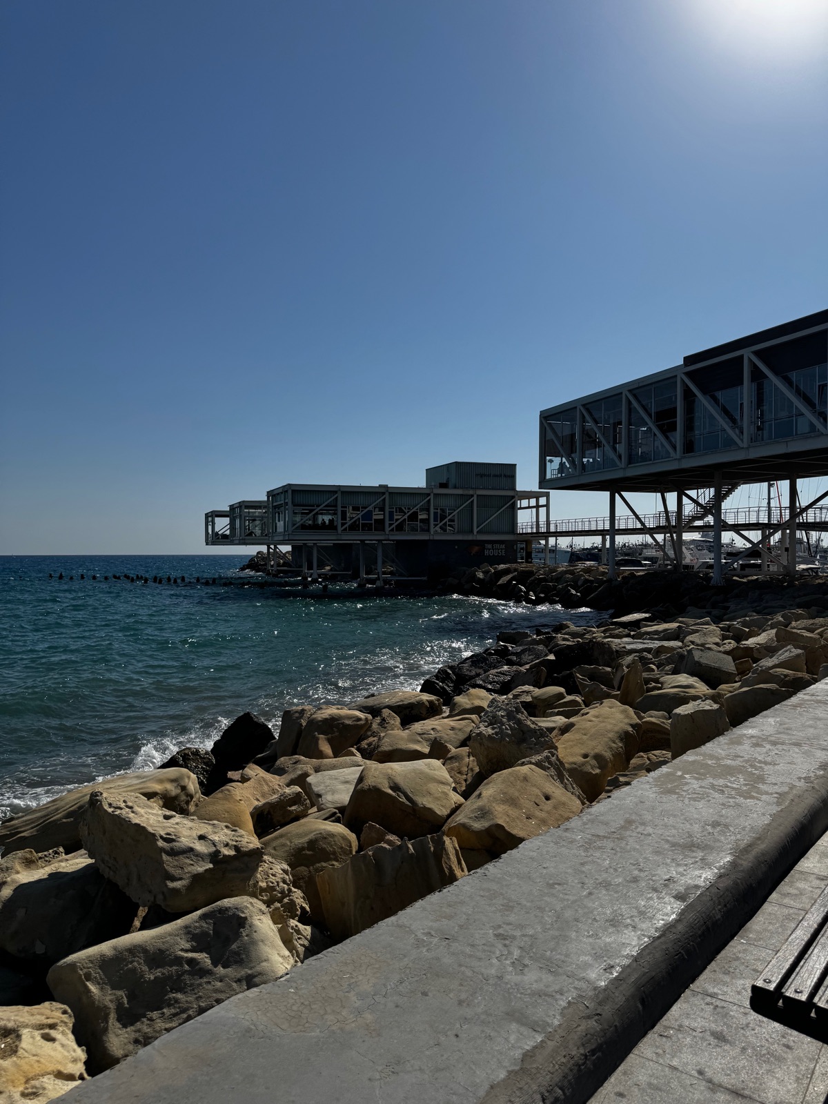 Γυάλινα κτίρια εστιατορίων να στέκονται πάνω σε πασσάλους πάνω από βραχώδη κυματοθραύστη δίπλα στο παλιό λιμάνι της Λεμεσού, σκούρα μπλε θάλασσα μπροστά