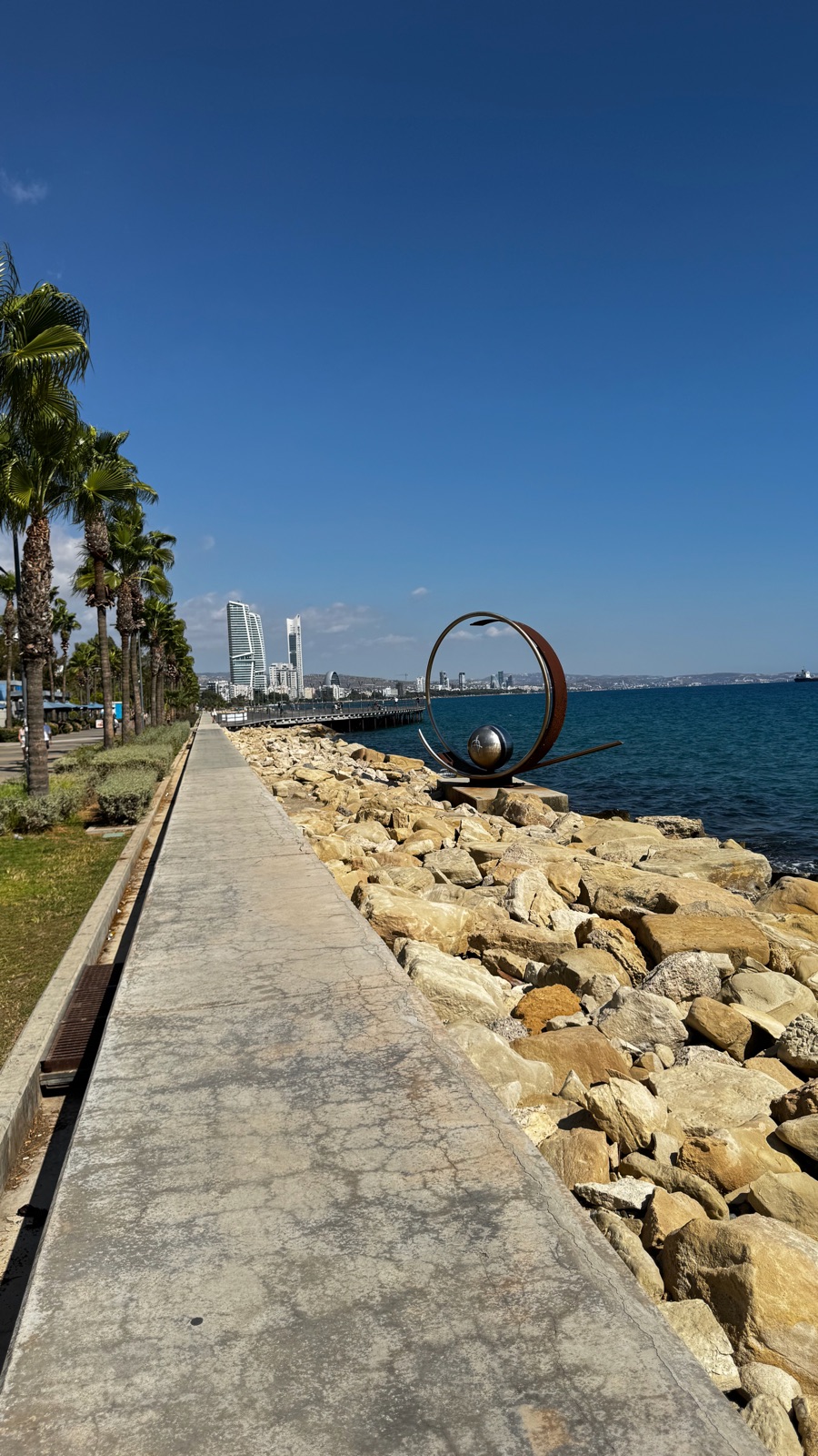 Ένα μεγάλο ατσάλινο γλυπτό-δαχτυλίδι με σφαίρα να στέκεται στη βραχώδη ακτή της Λεμεσού, ο περίπατος και η μακρινή γραμμή ορίζοντα πίσω